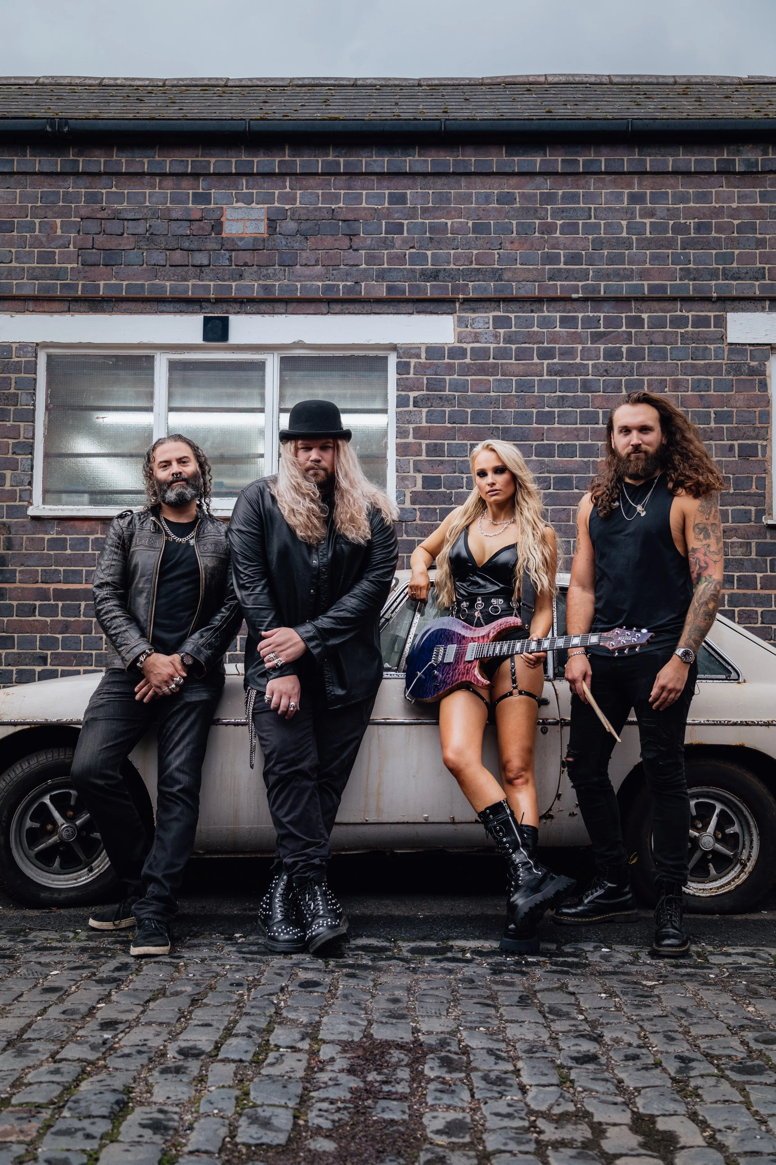 Four musicians standing in front of a vintage car, against a brick building. The group includes a man with long curly hair and a bushy beard, a man with long blonde hair and a black hat, a woman with long blonde hair holding a guitar, and a man with 
