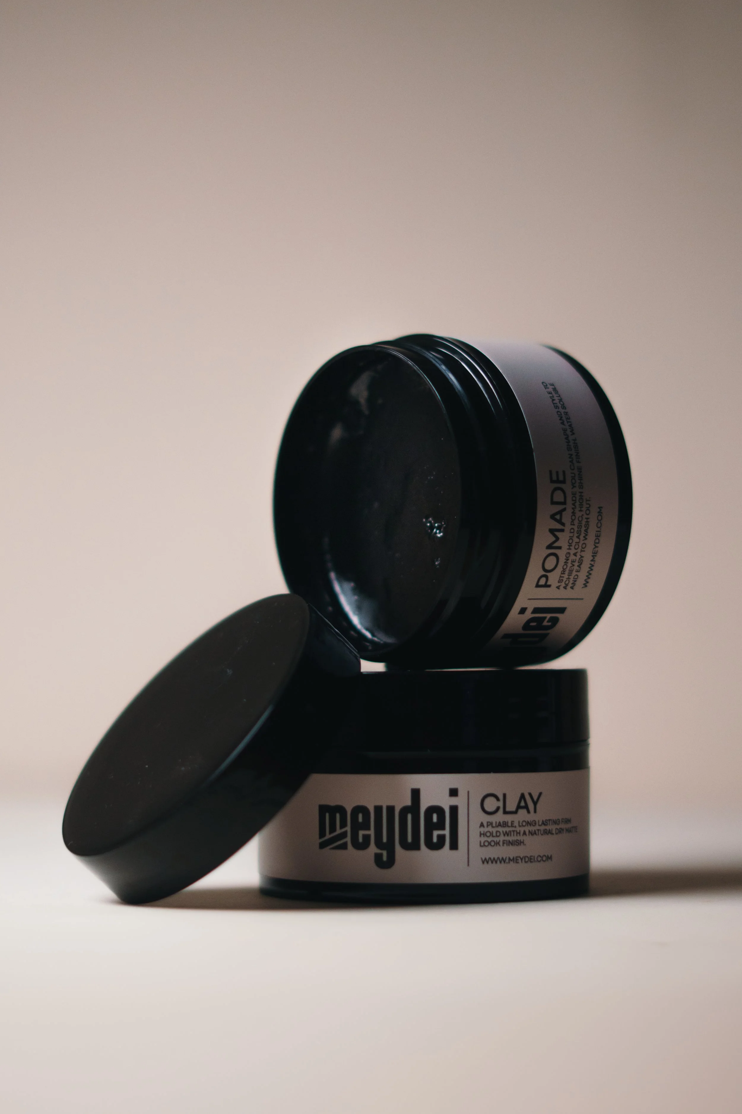 Two black containers of Meydeli clay, one open and upside down, on a plain surface with a neutral background.