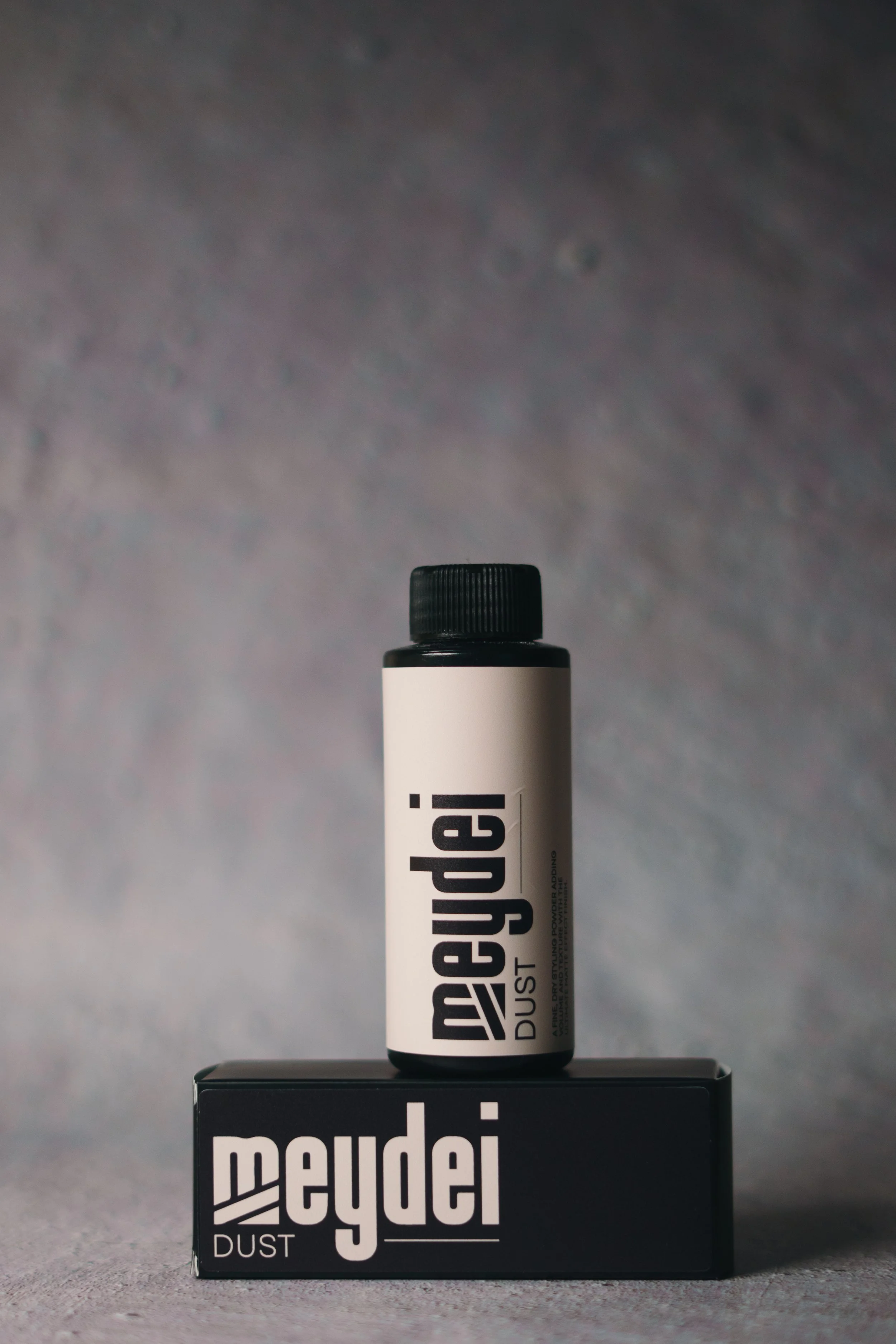 A black and white container of Meydei Dust powder placed on top of a matching black box, both featuring the Meydei logo, against a gray background.