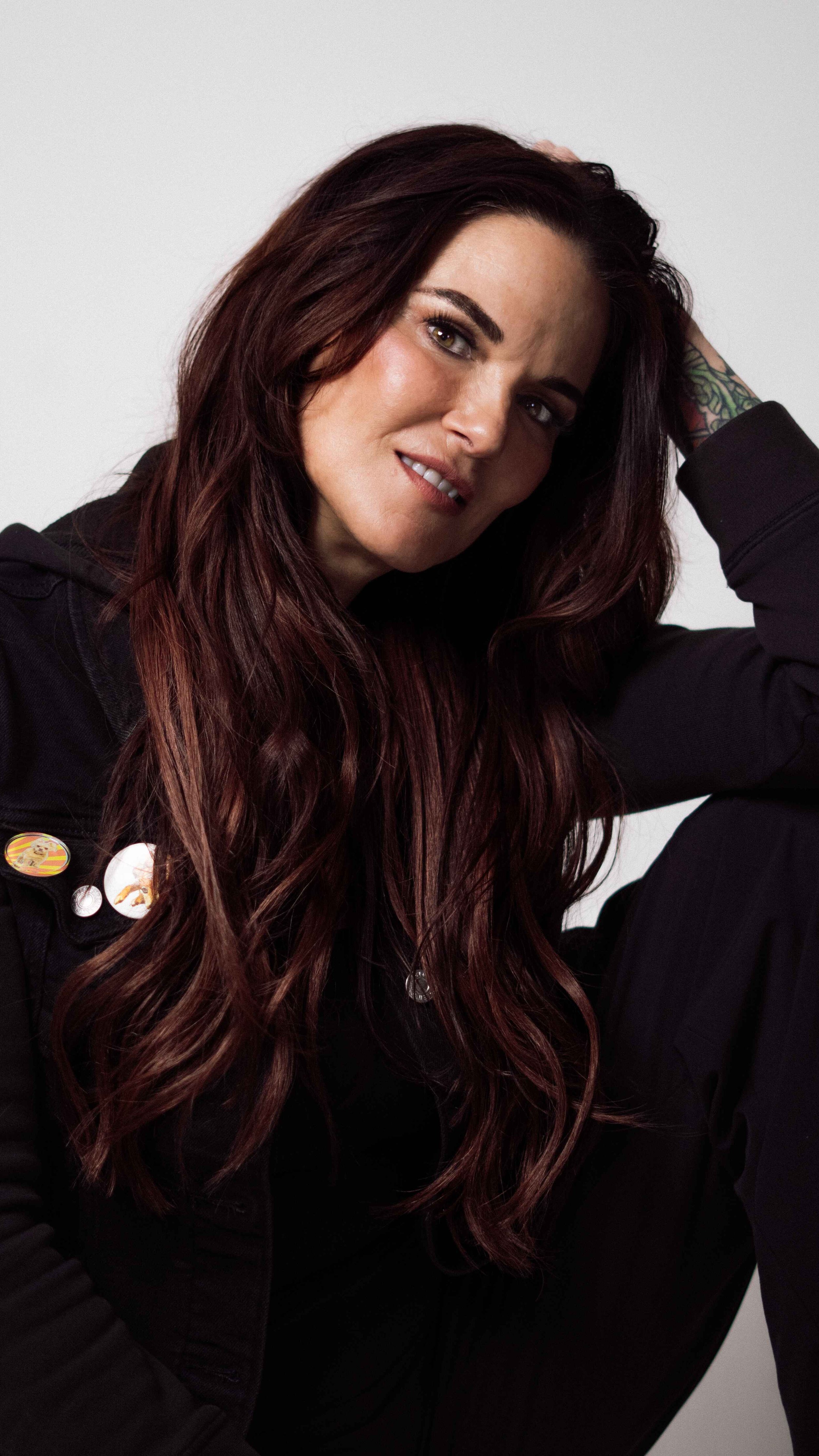 A woman with long, wavy, dark red hair, wearing a black jacket with some pins, sitting against a plain white background, resting her head on her hand.