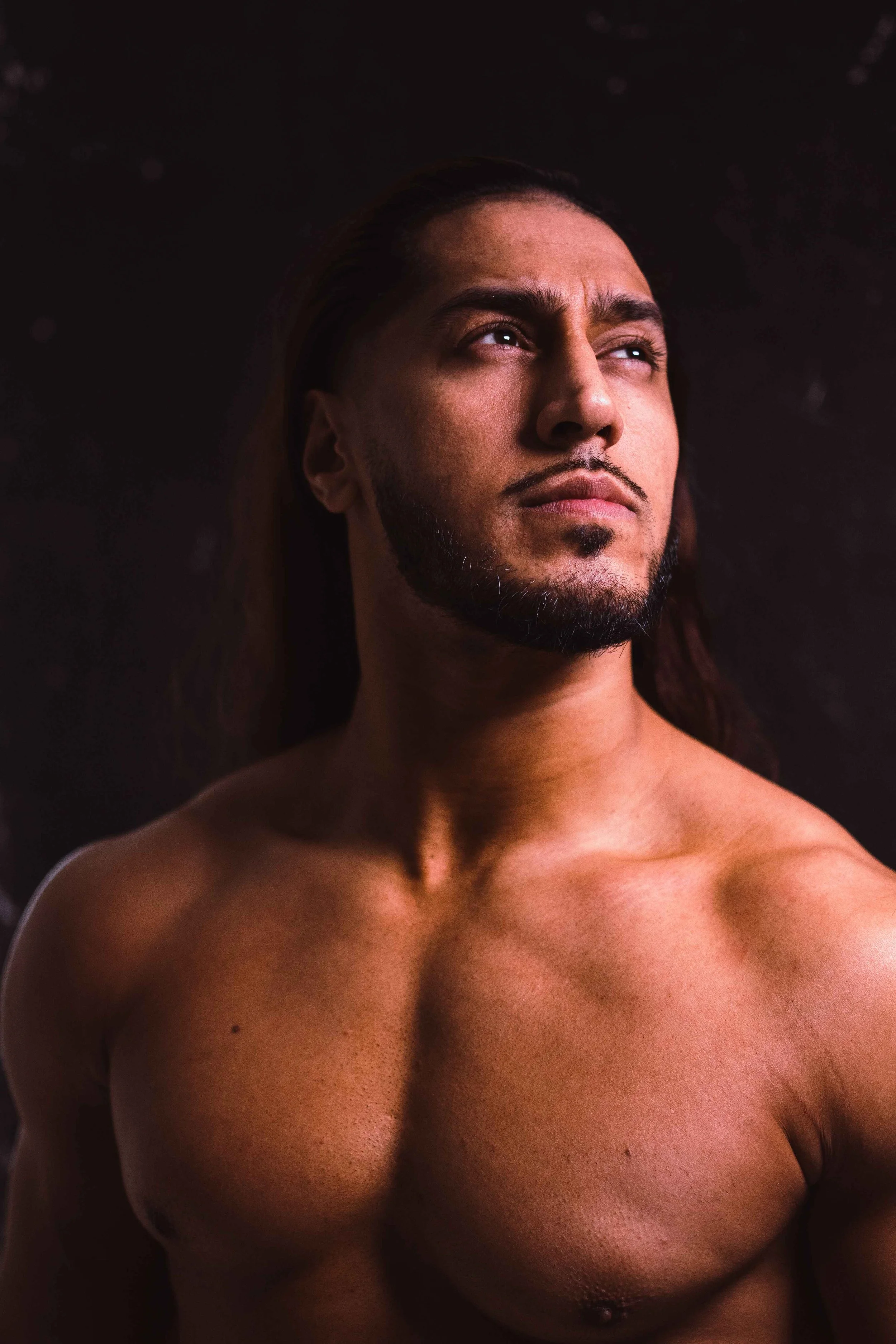 A shirtless man with long dark hair and a styled beard gazing upward against a dark background.
