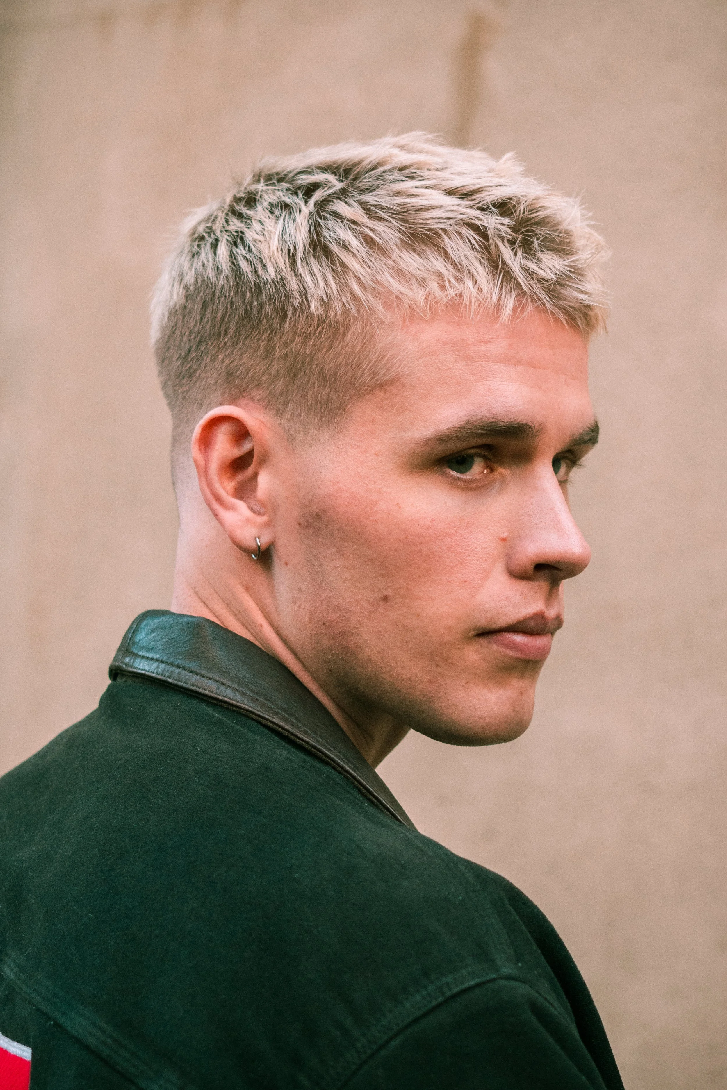 Close-up of a young person with platinum blonde short hair, wearing a black jacket with a leather collar, and a small hoop earring in the left ear, looking over their shoulder.