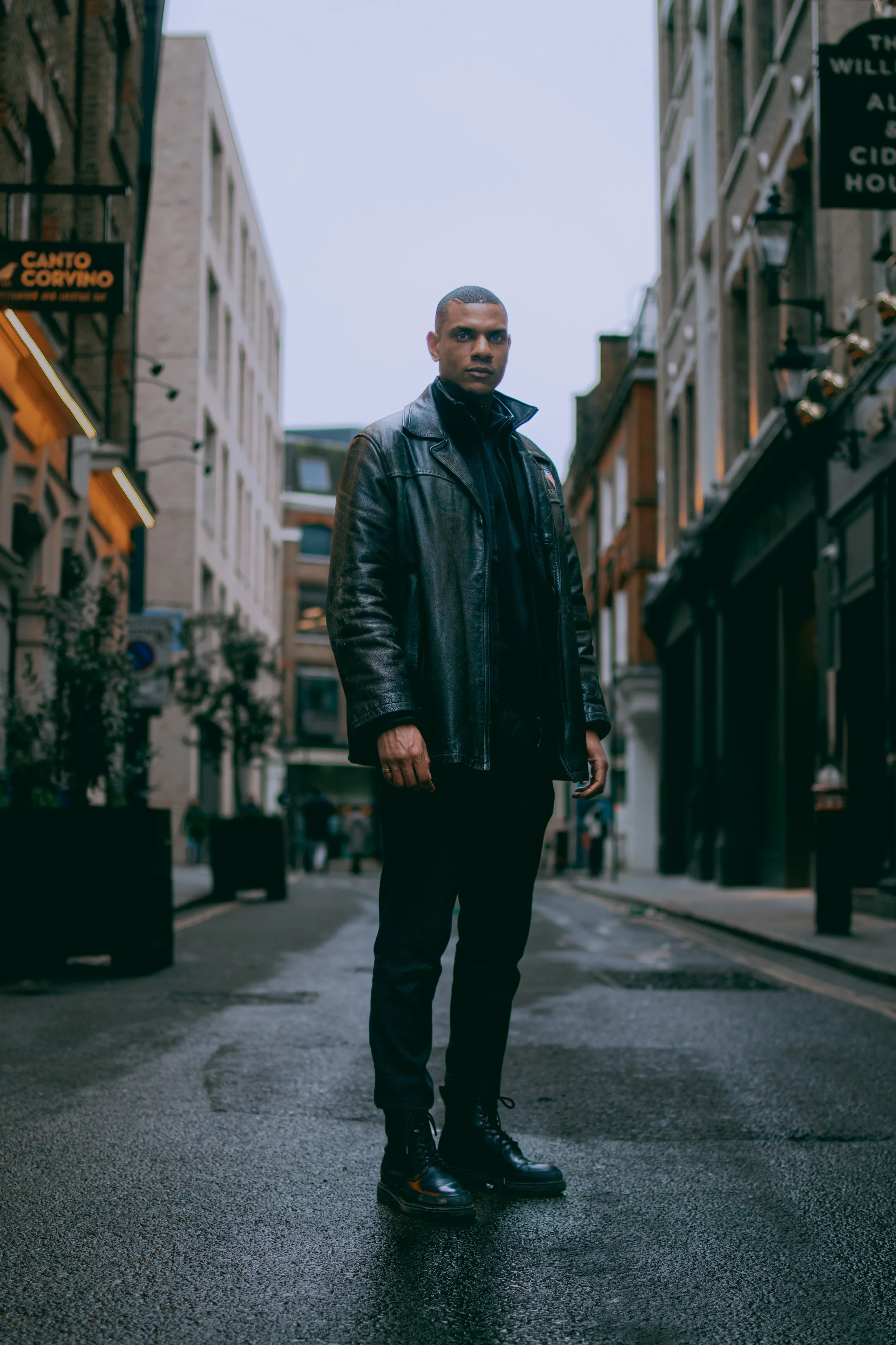 A man dressed in black standing on a wet city street at dusk, with buildings and signs visible around and a cloudy sky overhead.