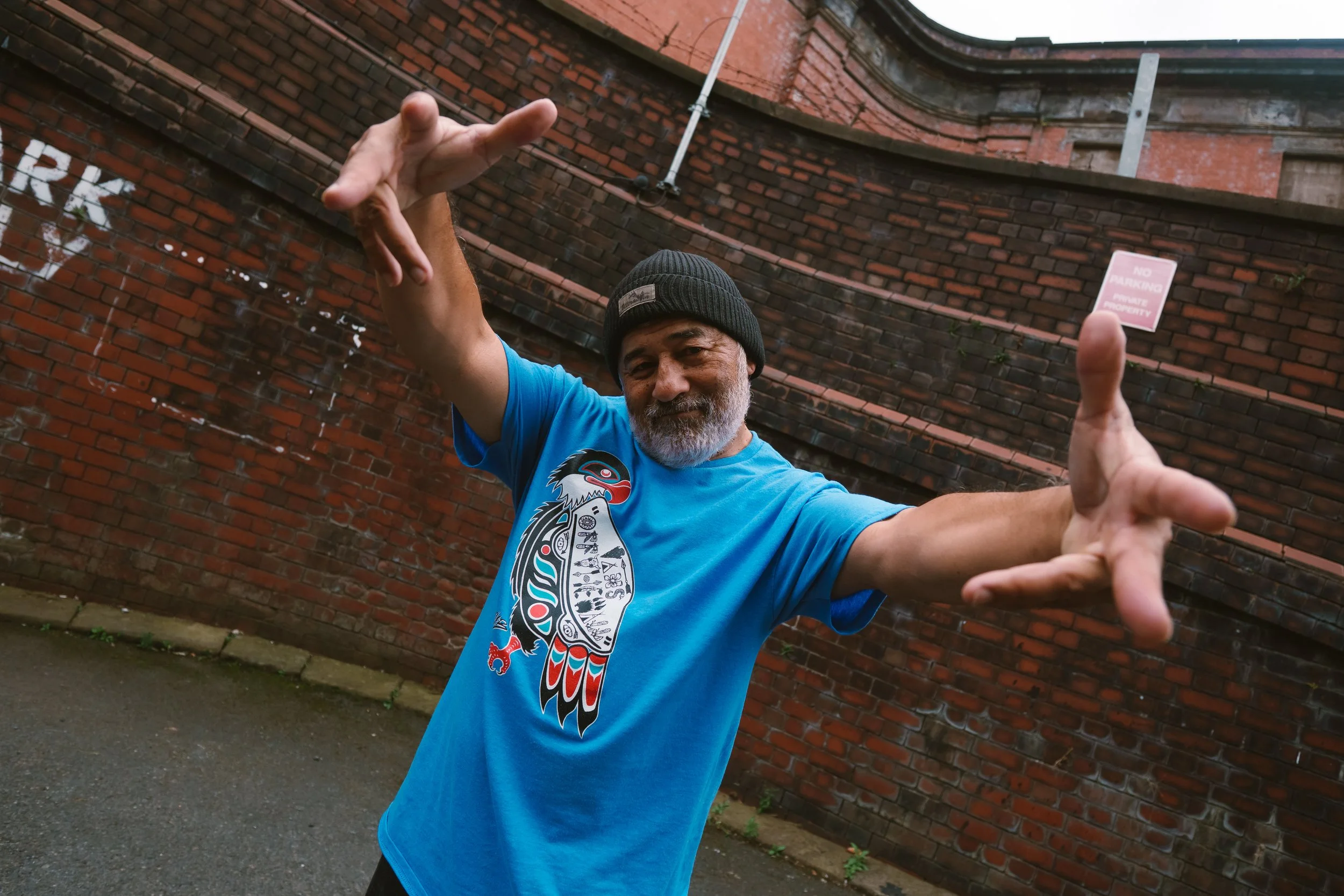 An older man with gray beard and mustache, wearing a black beanie and a bright blue T-shirt with a graphic design, is posing with outstretched arms in front of a red brick wall.