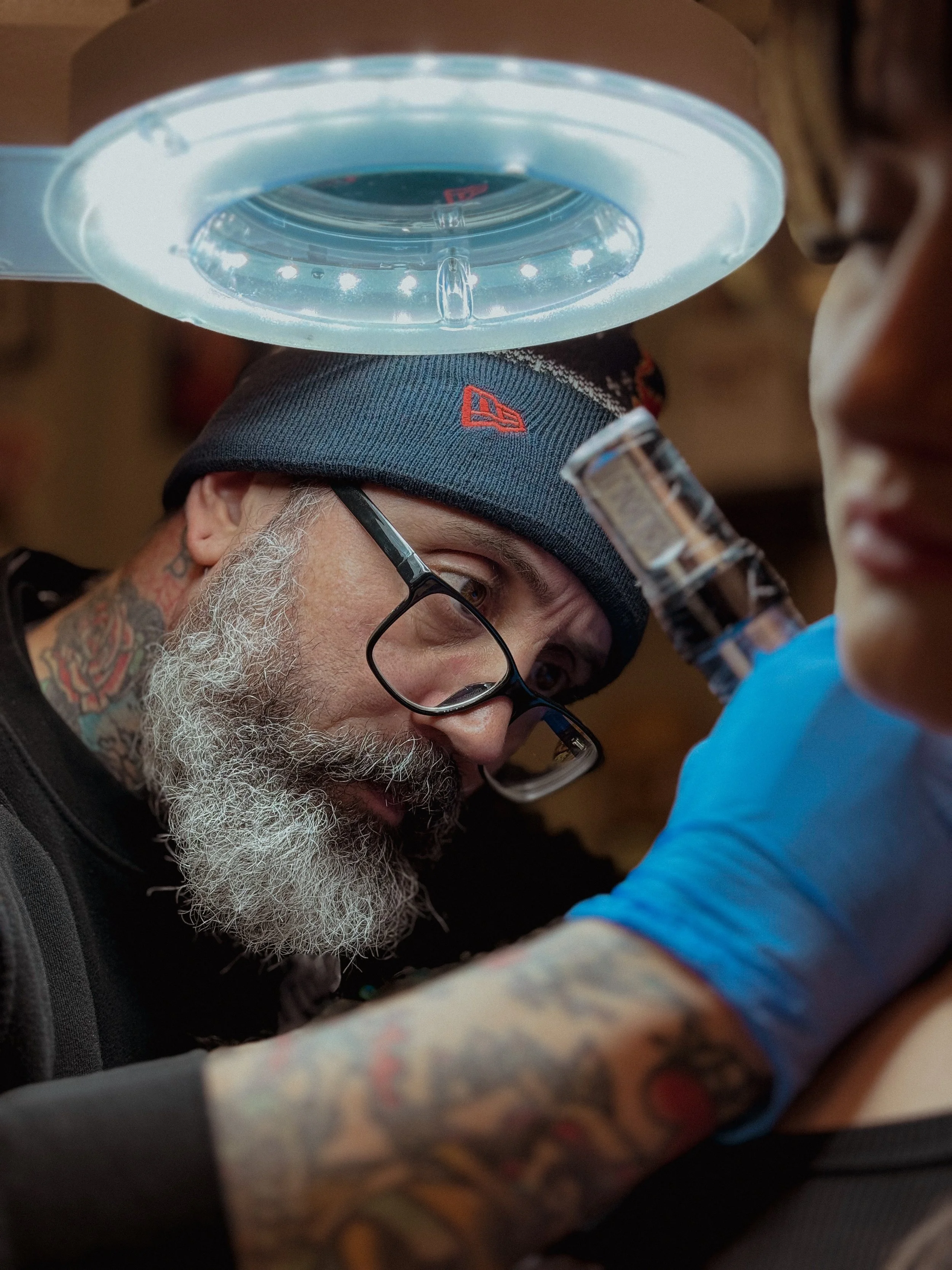 A tattooed man with glasses and a beanie performing tattooing on a client's arm with a tattoo machine, under a magnifying light.
