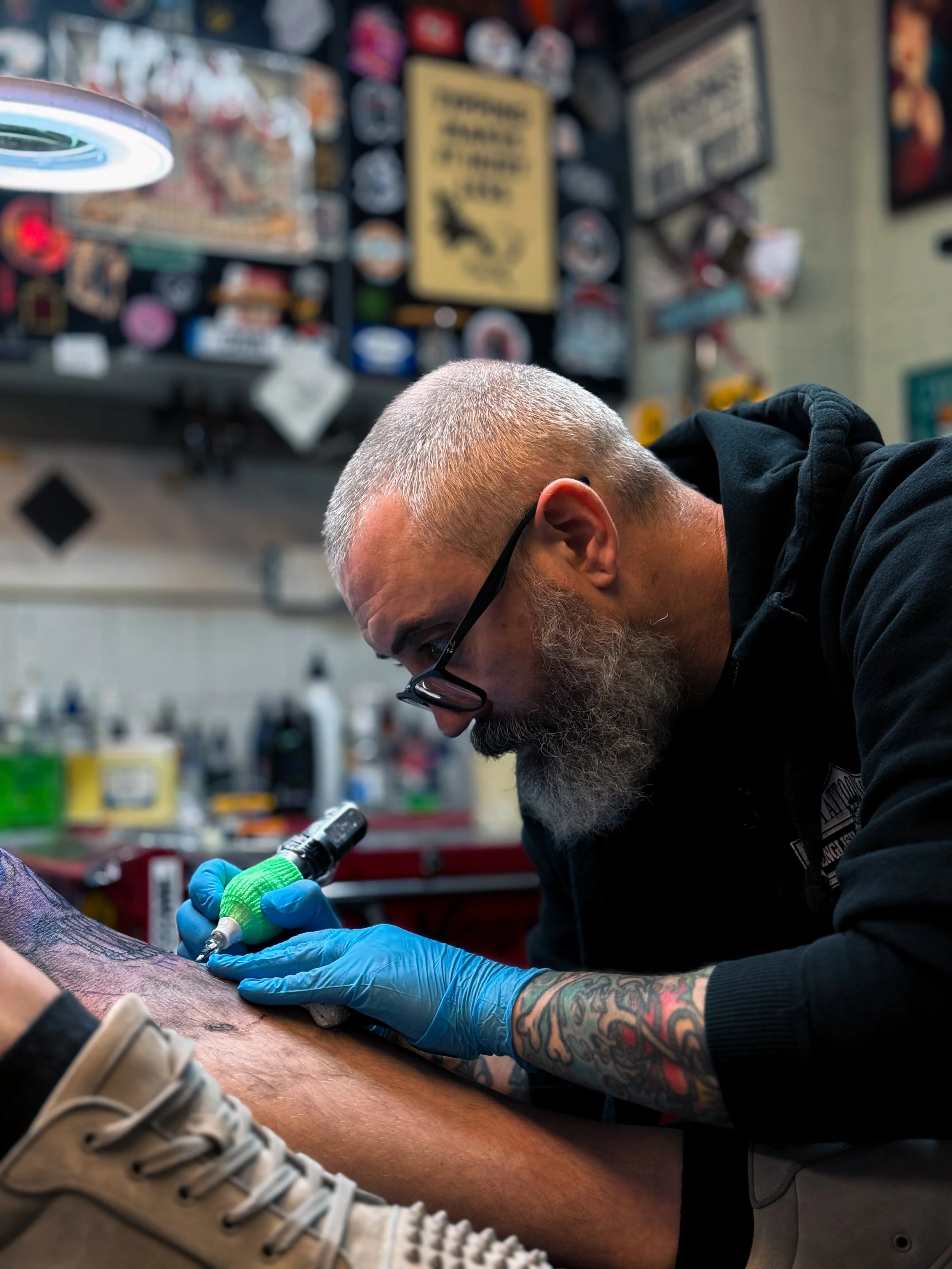 A tattoo artist with a gray beard, glasses, and tattoos on his arm, wearing a black hoodie and blue gloves, is tattooing a person's leg in a tattoo shop. The shop is decorated with various signs and artwork on the wall in the background.
