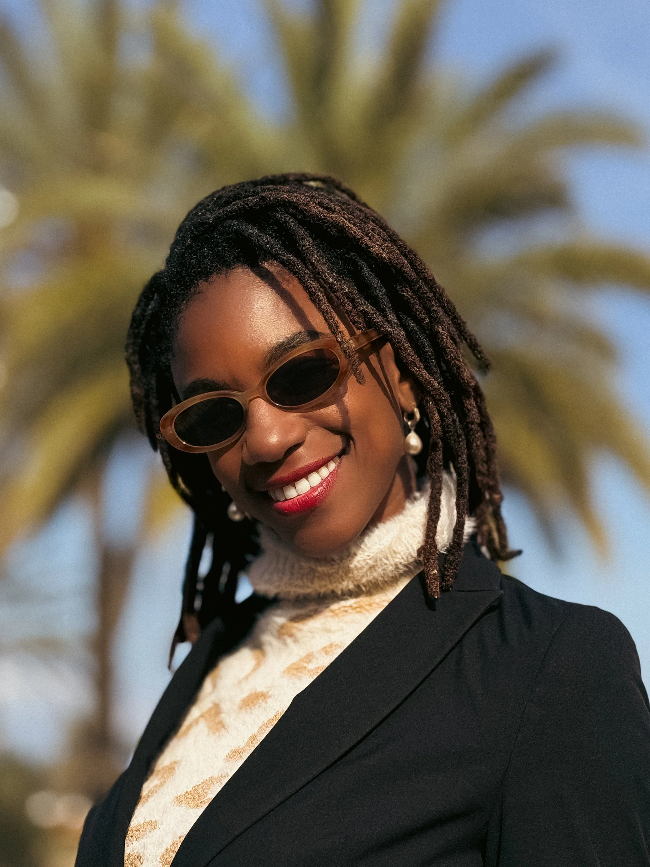 Smiling woman with dreadlocks wearing sunglasses, earrings, black blazer, and a cream turtleneck sweater outdoors with a palm tree in the background.