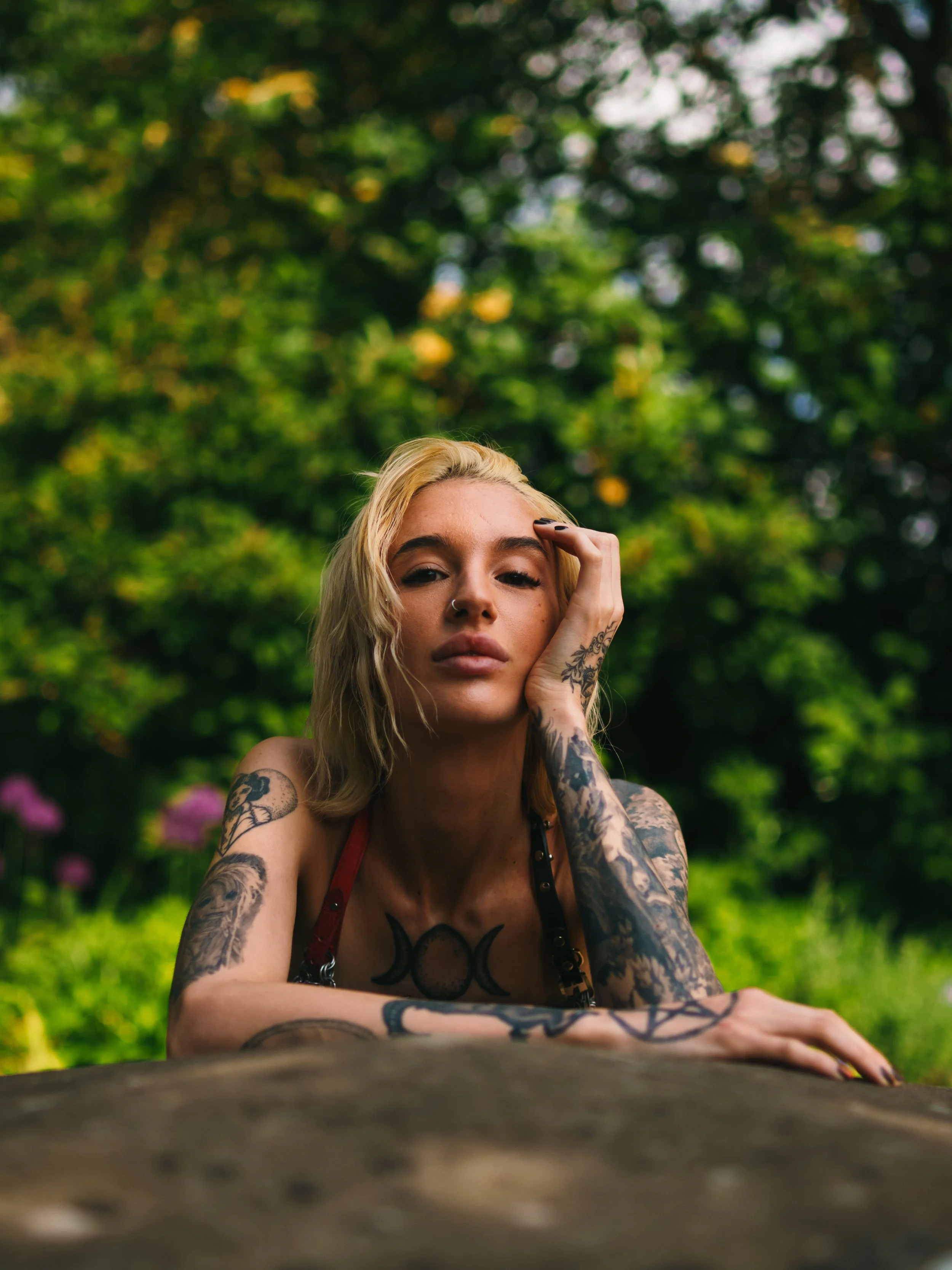 A woman with blonde hair and tattoos resting her face on her hand outdoors, with a lush green background and pink flowers.