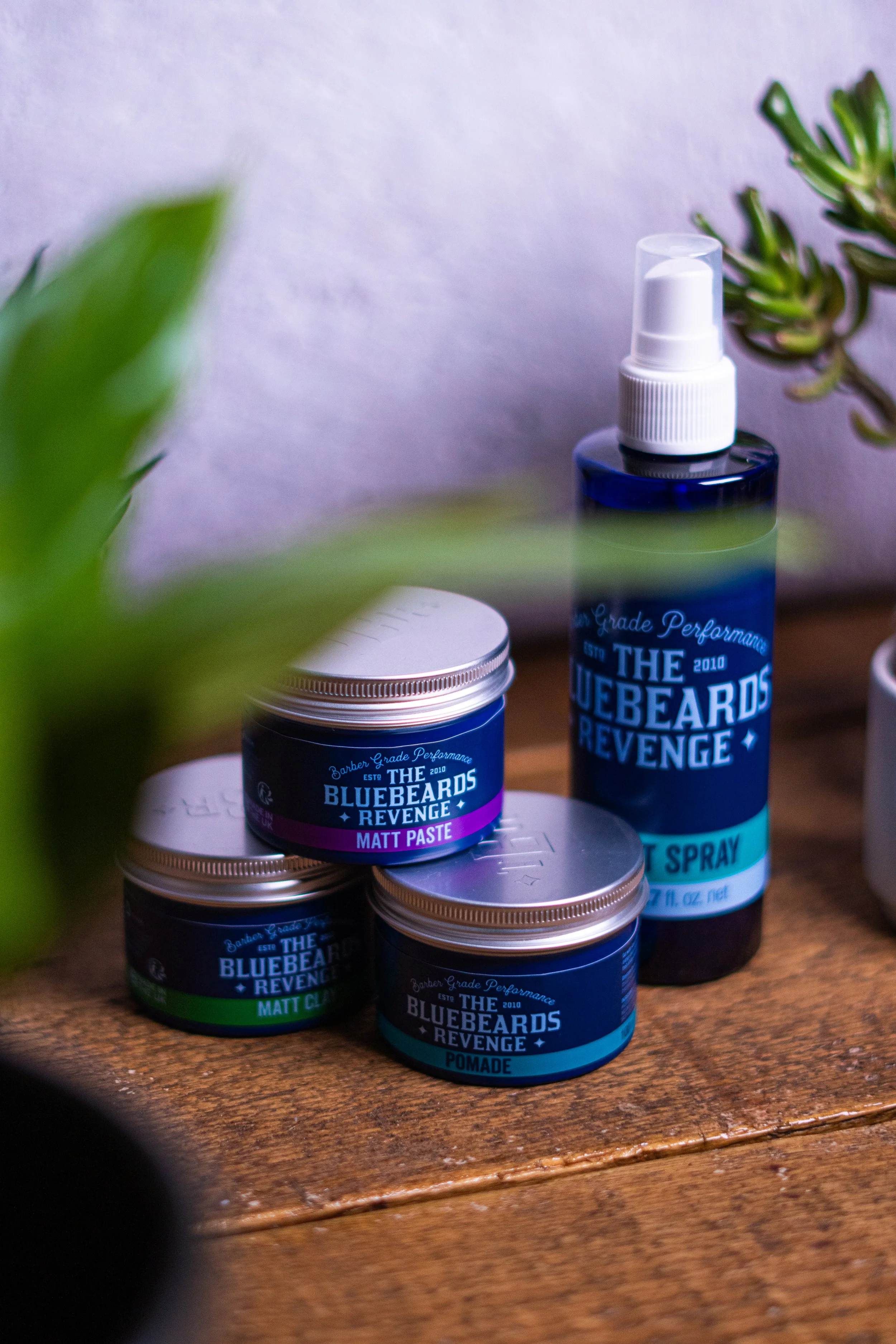 Hair styling products including pomade, matt paste, and hair spray on a wooden surface with plants nearby.