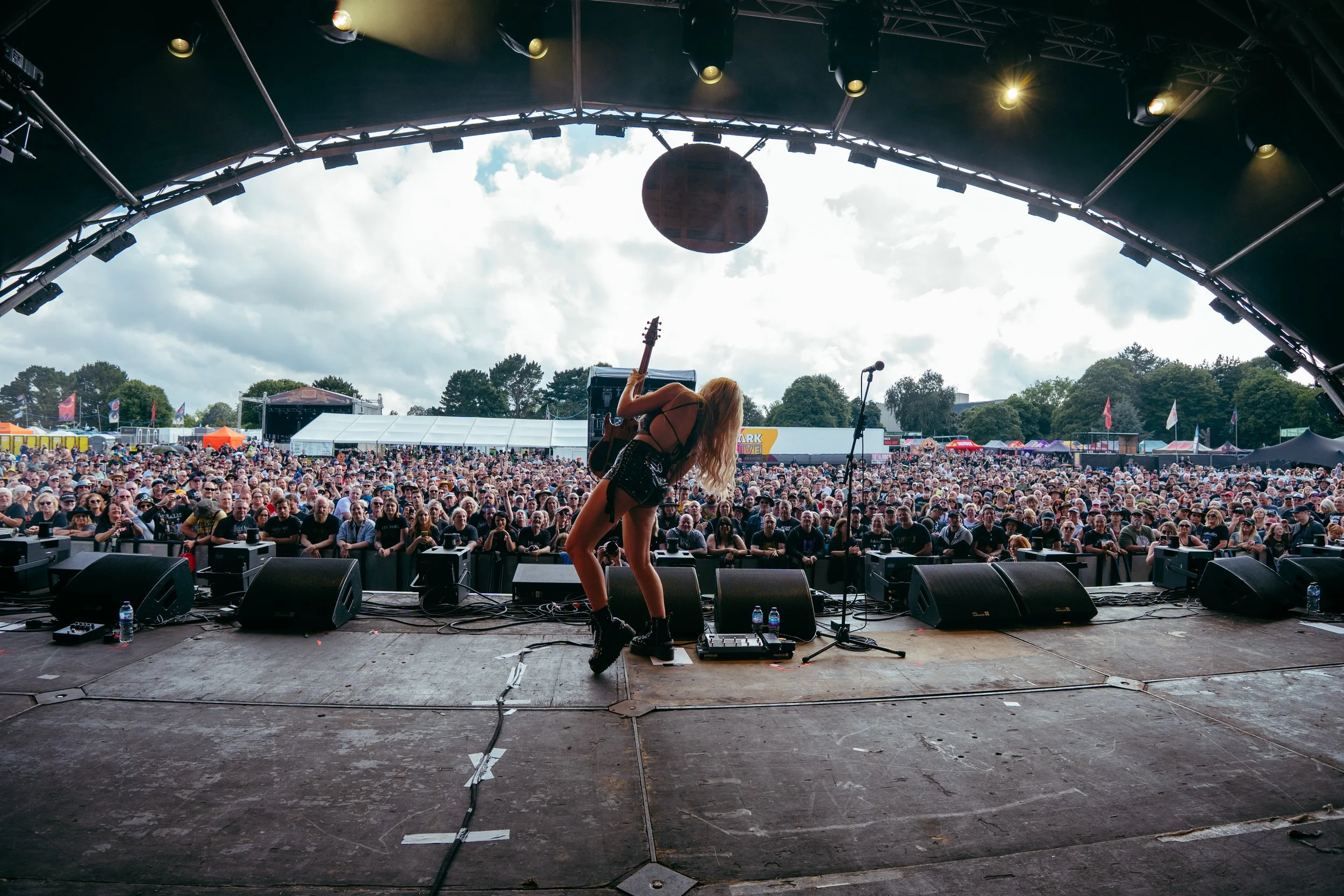 Female musician on stage playing guitar at an outdoor music festival with a large crowd watching.