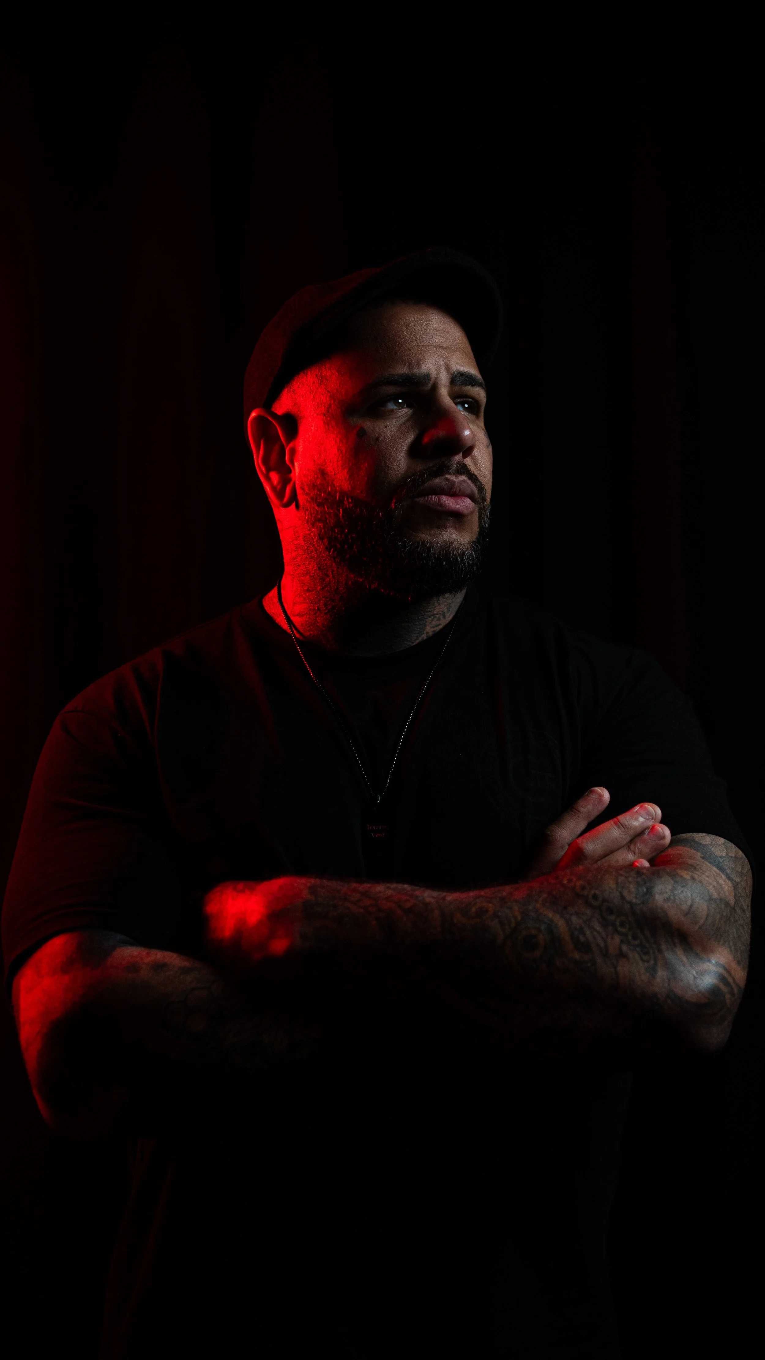 A man with tattoos on his arms, wearing a black shirt and a necklace, stands with arms crossed against a dark background. Red lighting illuminates part of his face and arm.