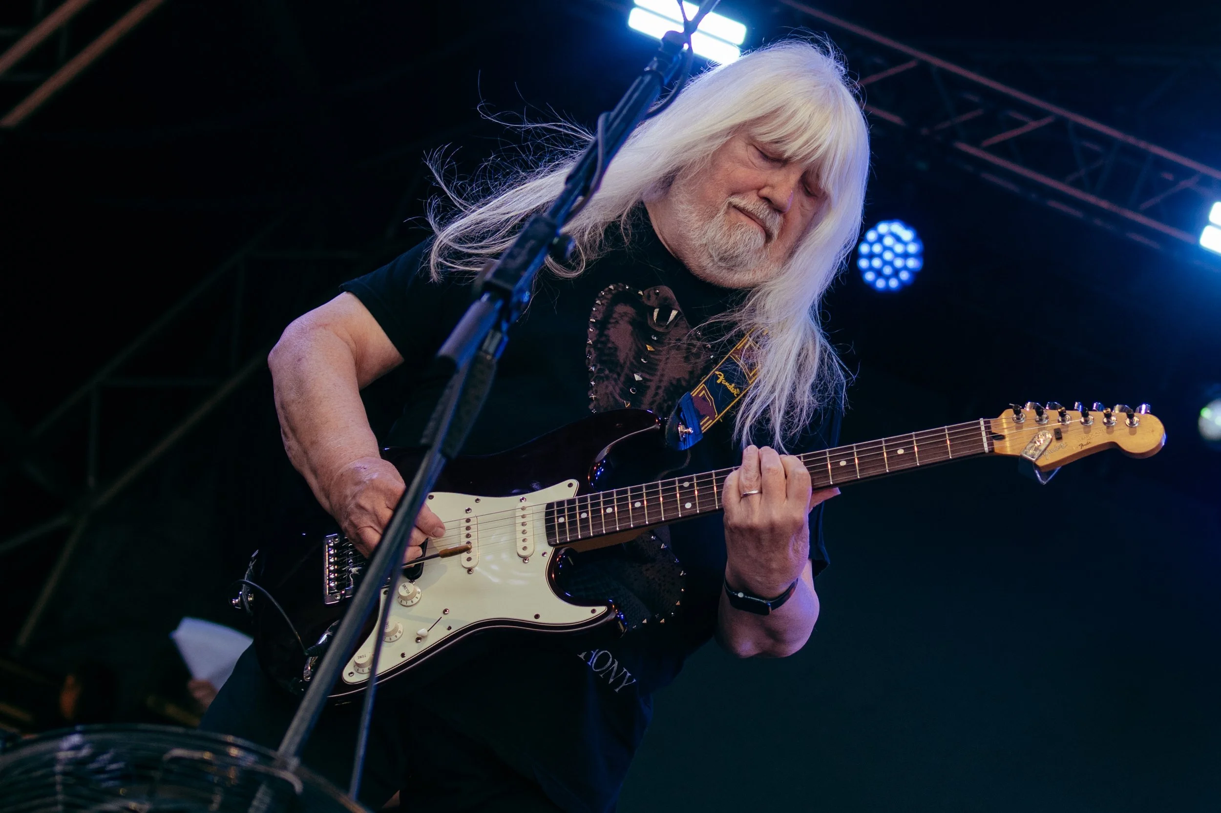 An elderly man with long white hair and beard playing an electric guitar on stage under blue and white stage lights.