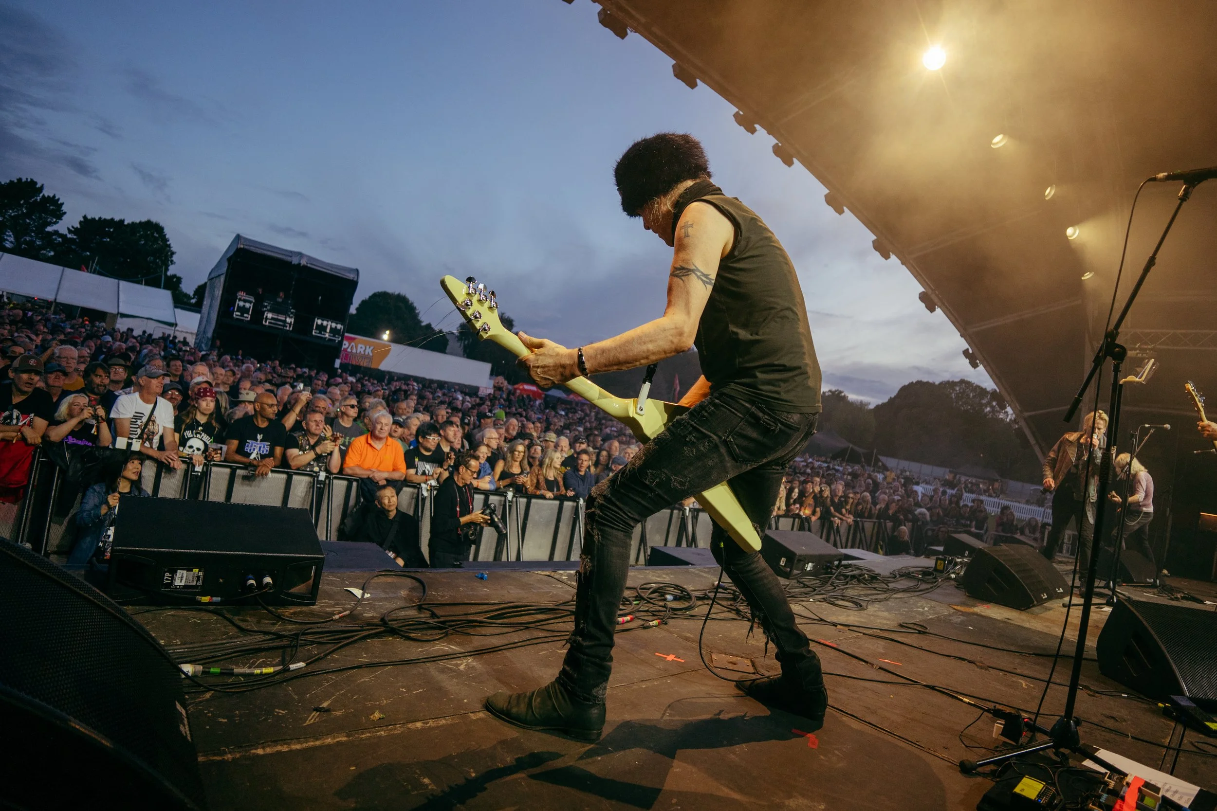 A musician with tattoos playing an electric guitar on stage during an outdoor concert at dusk, with a large audience watching.