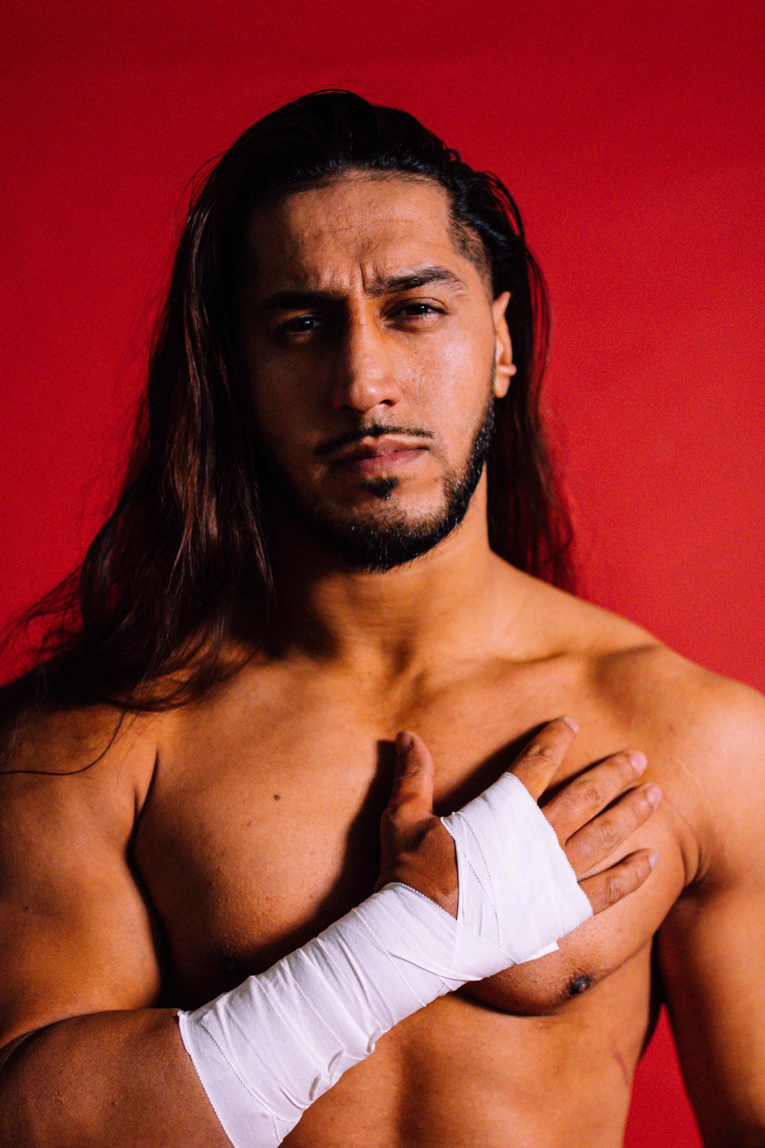 A shirtless man with long hair and a beard, covering his heart with his right hand wrapped in white bandages, against a red background.
