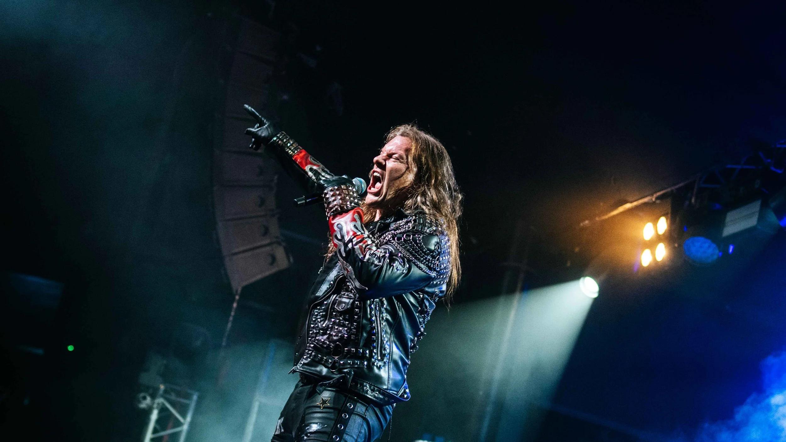 Rock singer performing on stage, wearing a black leather jacket with red and silver studs, holding a microphone, and singing passionately.