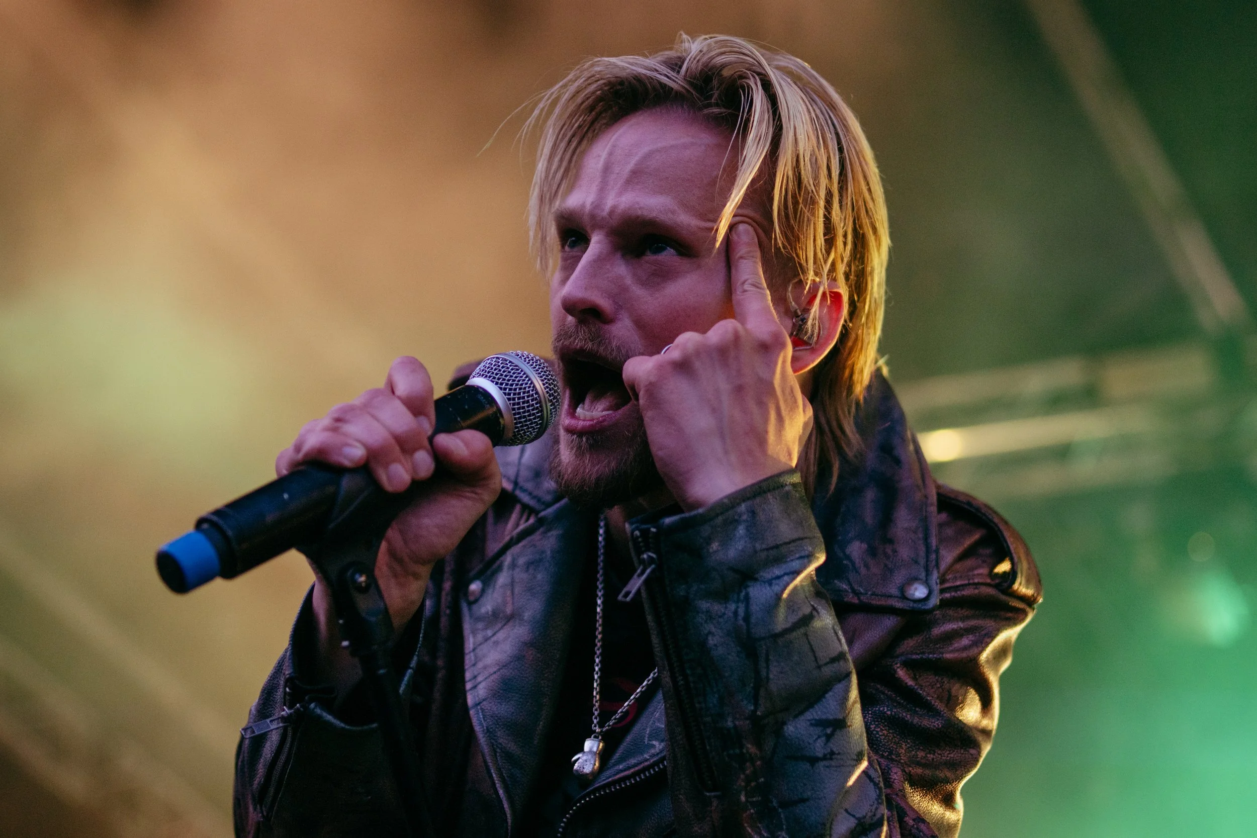 A male singer with blonde hair singing into a microphone, wearing a leather jacket, with his hand on his head, during a performance.