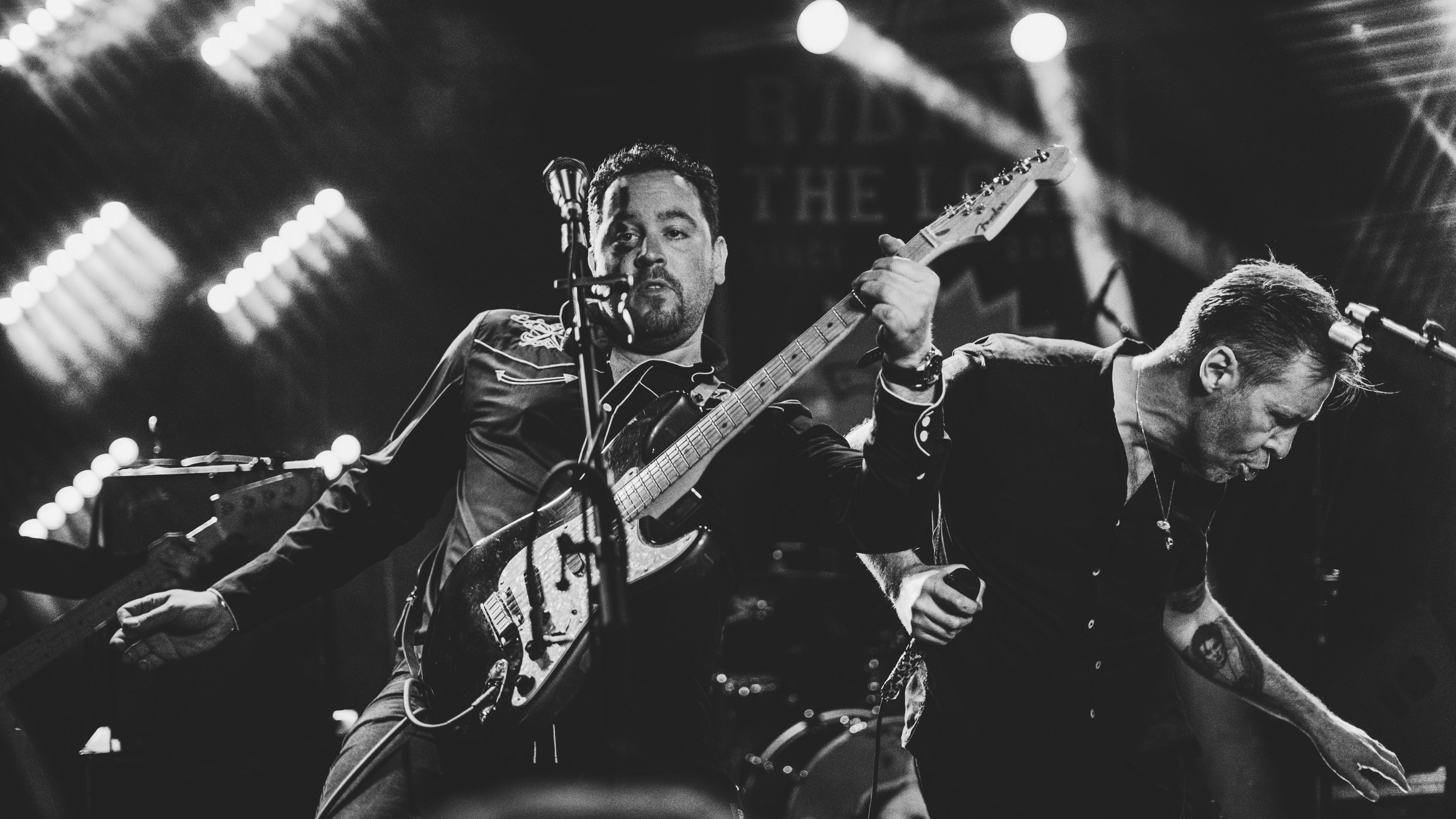 Black and white photo of two musicians performing on stage. One is playing an electric guitar and singing into a microphone, while the other is playing a keyboard. Stage lights create a bright background.