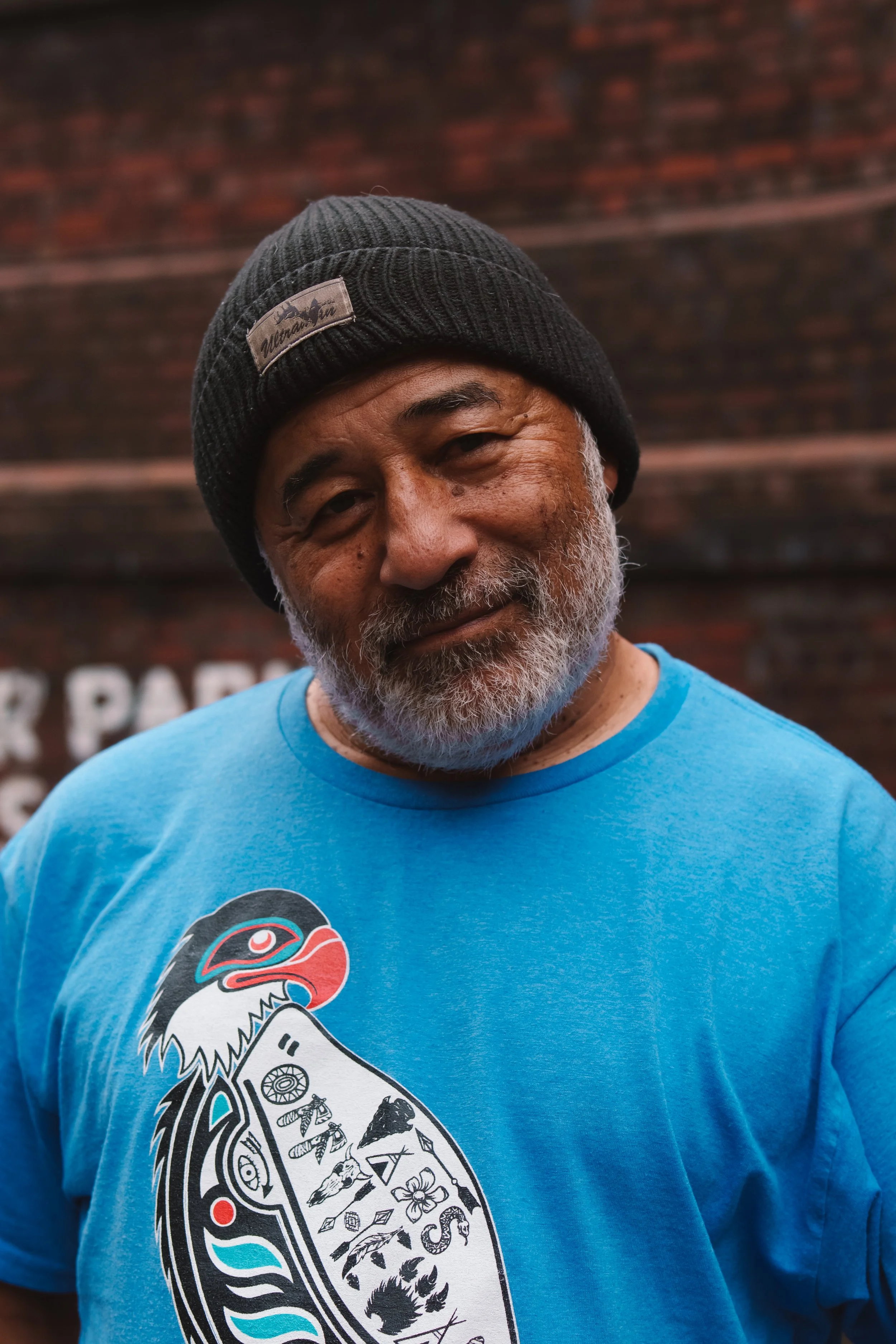 Close-up of an older man with a gray beard, wearing a black knit beanie and a blue T-shirt with a graphic of a bird and various symbols on it, standing outdoors against a blurred brick wall background.