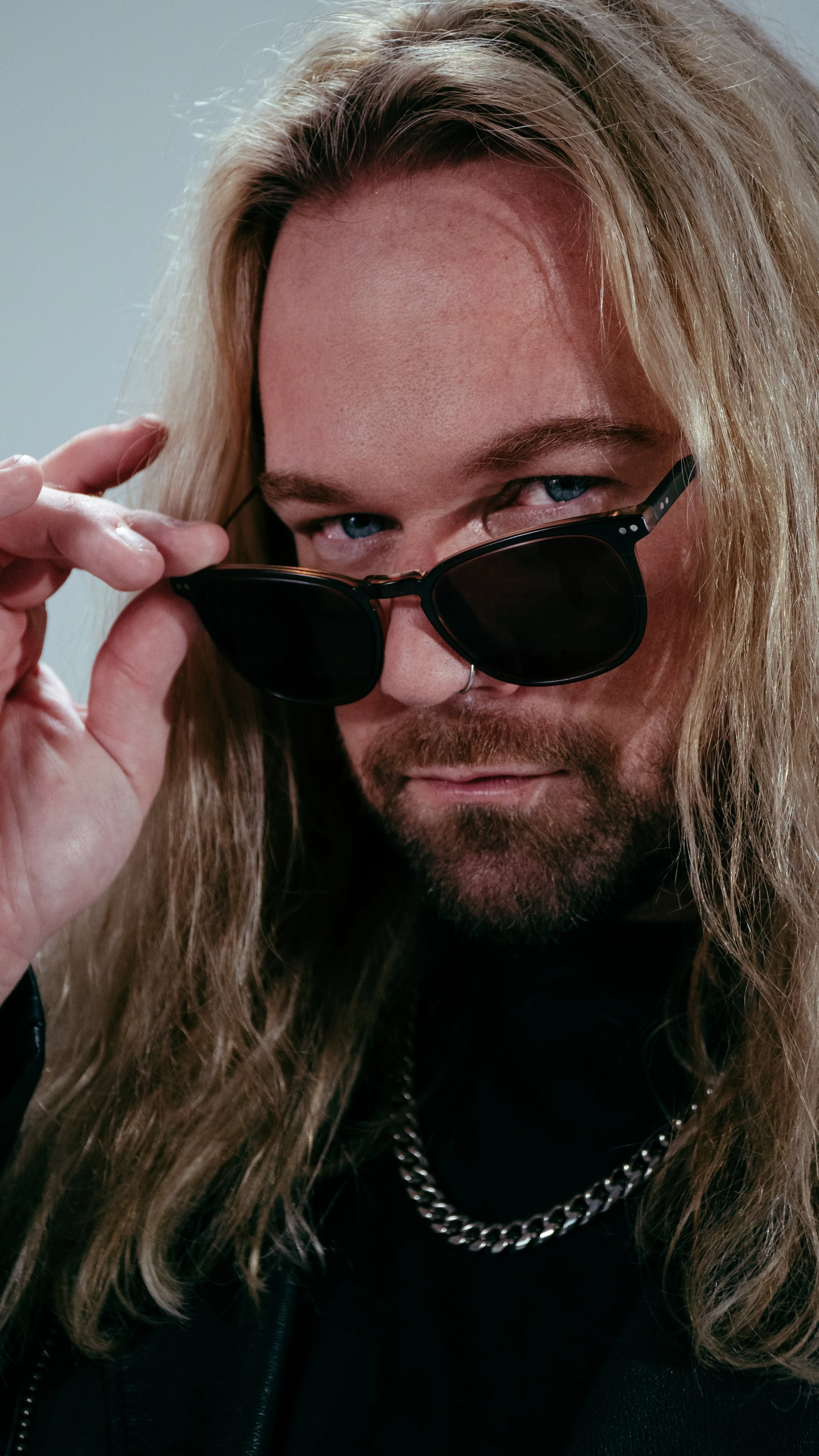 A man with long blonde hair and a beard lifting sunglasses to reveal blue eyes, wearing a black shirt and a silver chain necklace.
