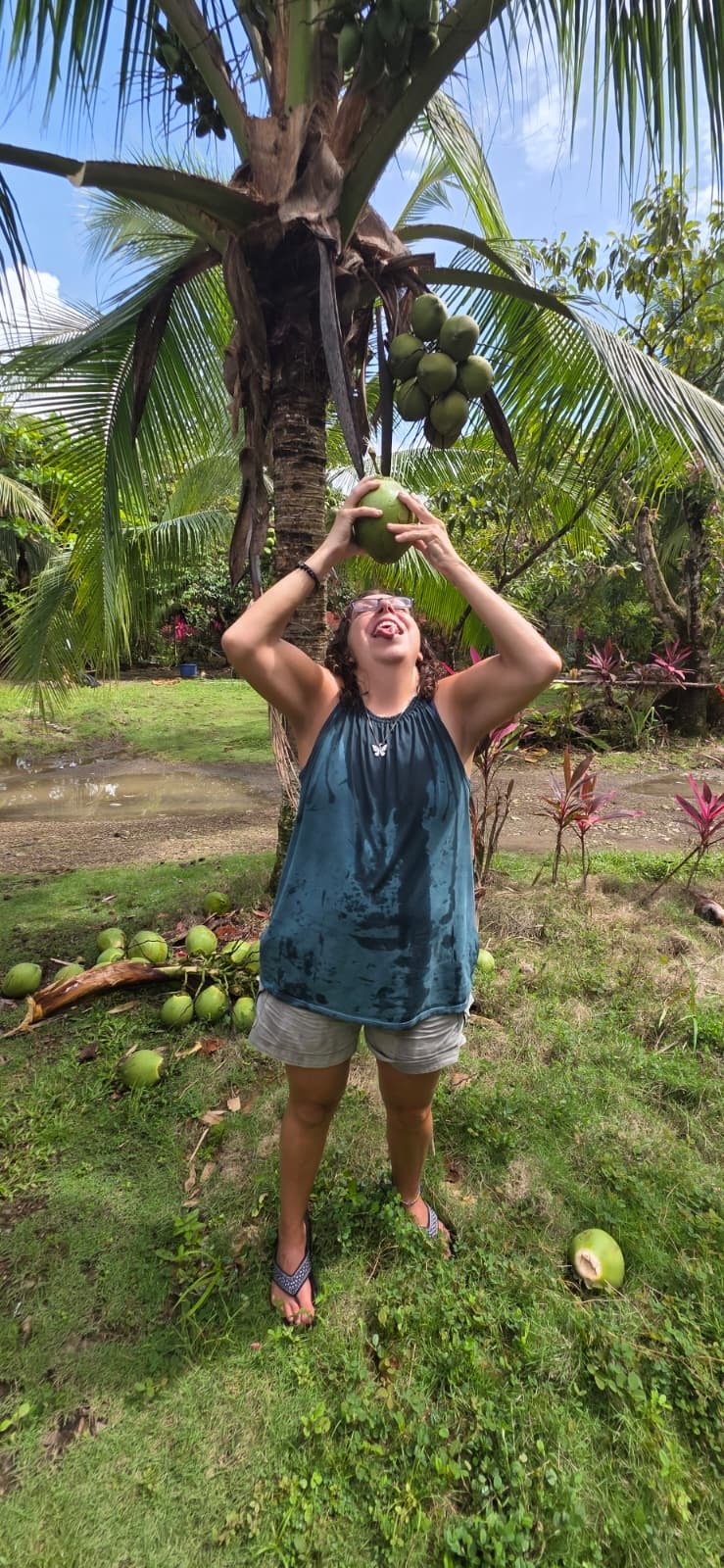 A woman stands under a coconut tree, holding a green coconut above her head with both hands, and sticking her tongue out playfully. The tree has multiple coconuts hanging from it, and there are more coconuts on the ground around her. She is wearing a wet, dark blue sleeveless top, beige shorts, and flip-flops, with a tropical garden and cloudy sky in the background.
