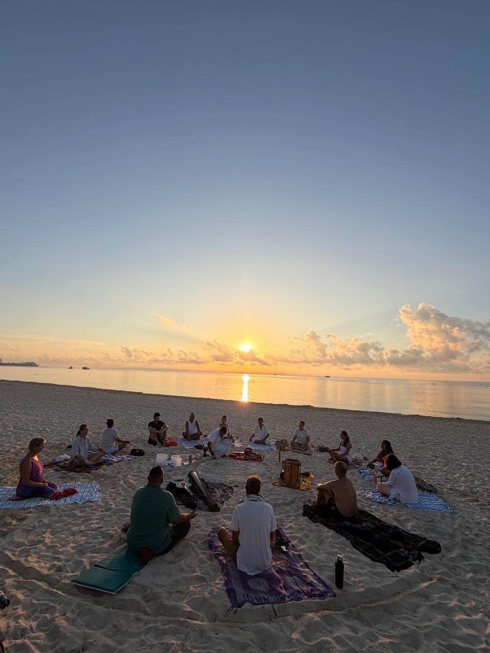 Group of people sitting on beach at sunset, participating in a meditation or yoga session on mats, facing the water.