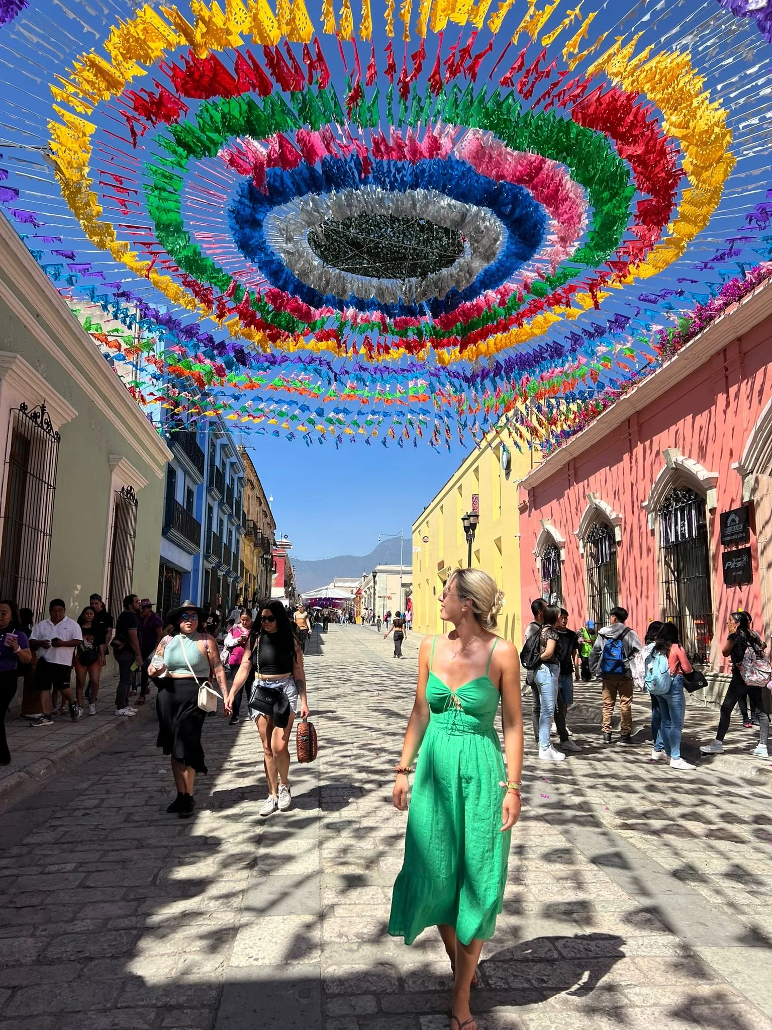 A crowded street scene during the day with colorful buildings and a large decorative canopy of rainbow-colored flags overhead. People are walking and socializing on the cobblestone street, with a woman in a green dress walking in the foreground.