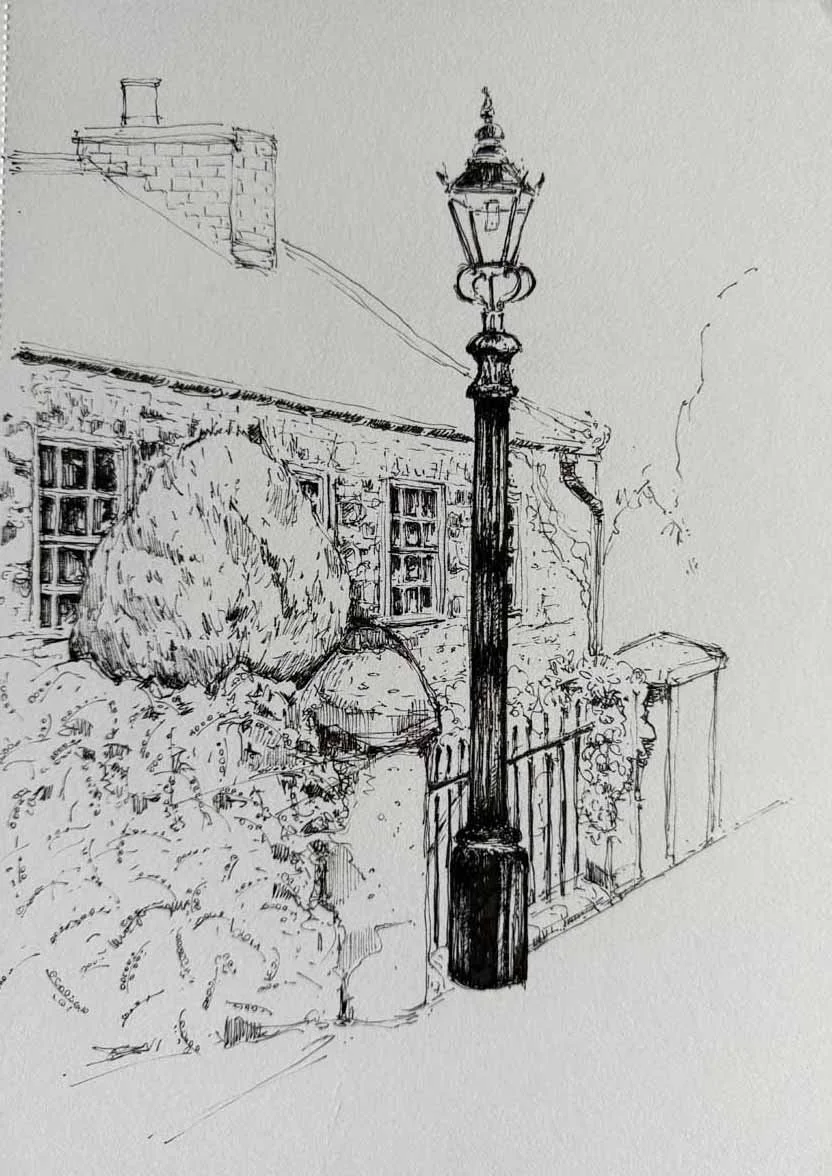 Line drawing of a street scene featuring a decorative lamp post, a house with brick chimney, and a garden with bushes and a fence.