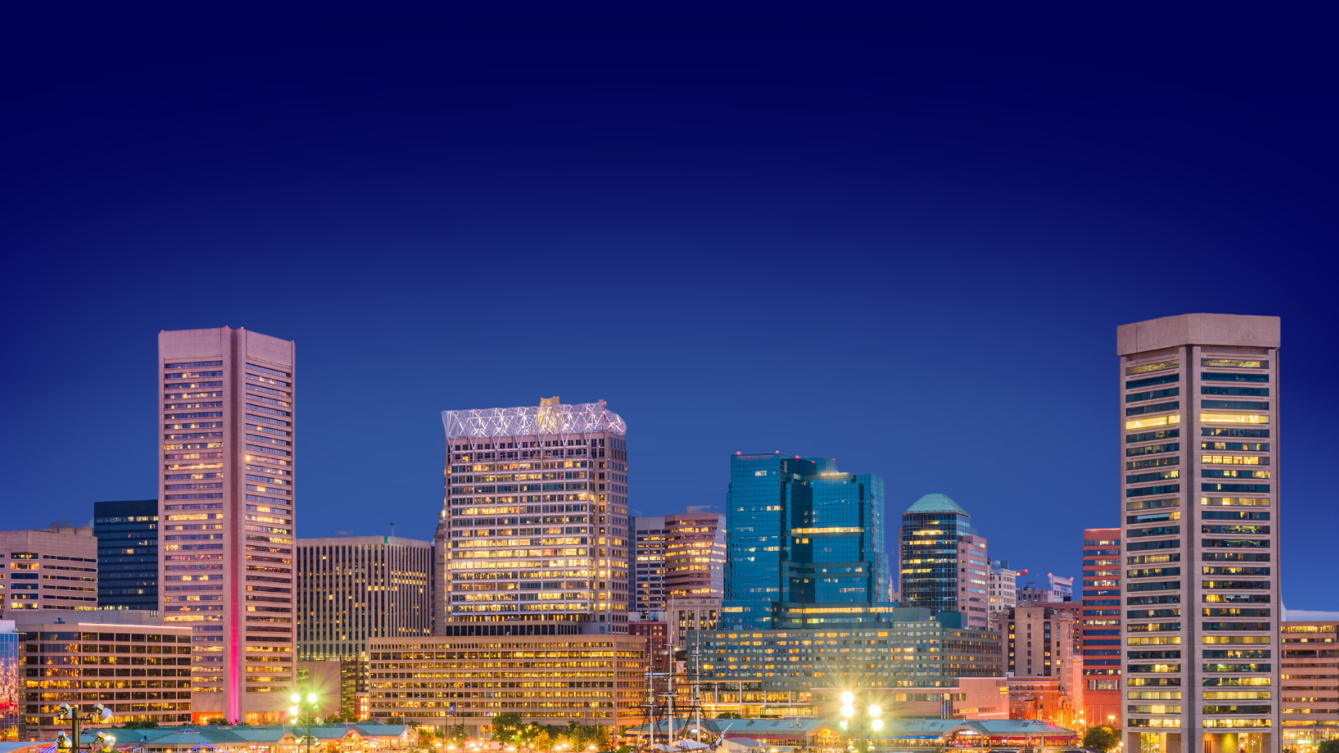 Nighttime city skyline with illuminated skyscrapers and buildings under a dark blue sky, some with lights on.