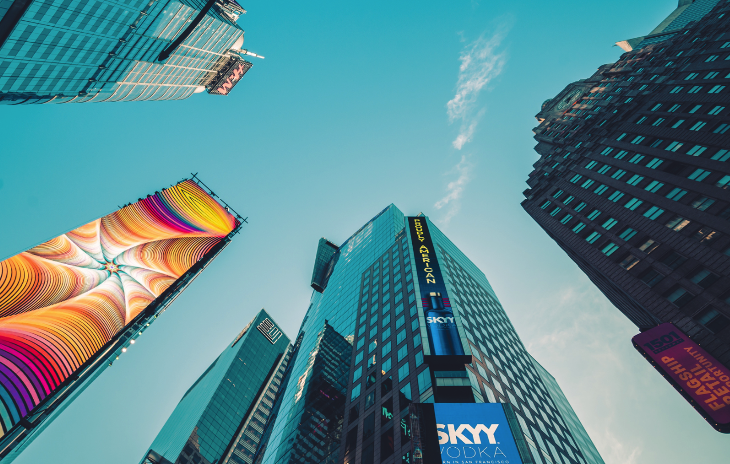 Looking up at skyscrapers in Times Square, New York City, with digital billboards and advertisements on the buildings against a blue sky.
