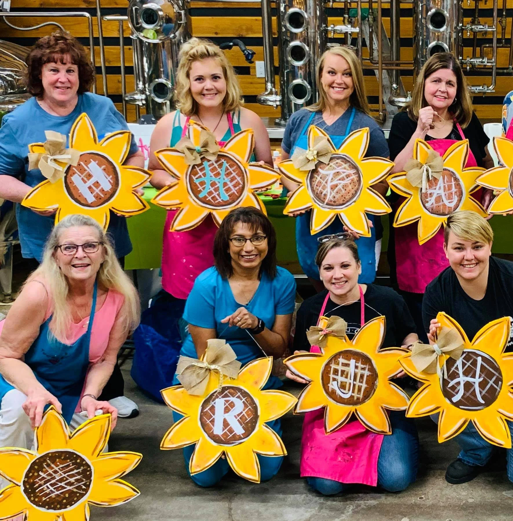 Group of women smiling and holding sunflower-shaped cookies with letter and island designs at a baking or craft event.