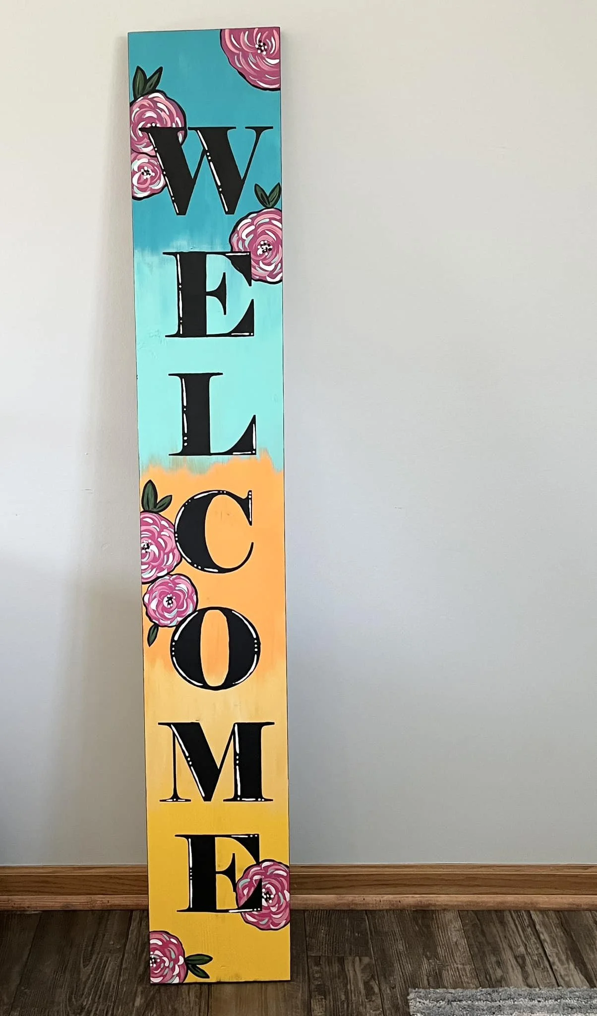 Vertical wooden sign with the word "WELCOME" written vertically in large black letters. The background is painted in a gradient of blue, orange, and yellow. Pink roses with green leaves are decorated around the letters.