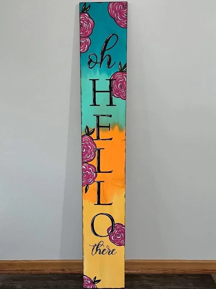 A tall, rectangular wooden sign painted with colorful gradients from teal to orange to yellow. The sign features the word "hello" vertically in large black letters, with the word "there" written in smaller cursive near the bottom. Pink floral designs