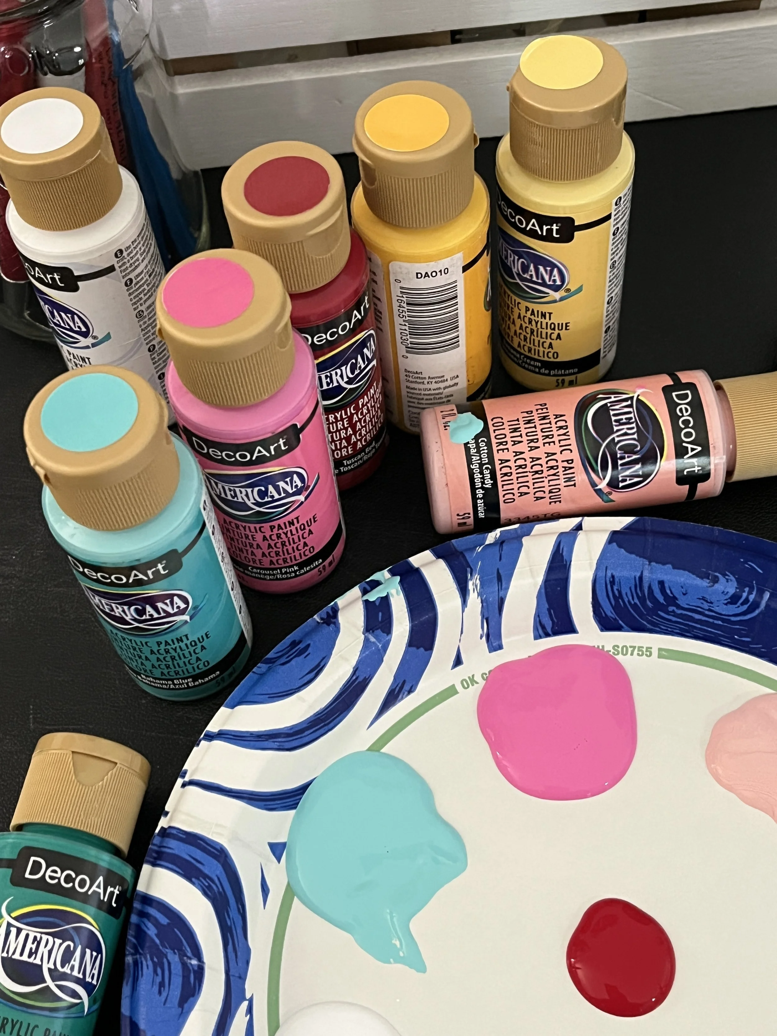 Assorted bottles of acrylic paint in various colors (white, red, pink, yellow, turquoise) placed on a black surface near a color palette with dollops of paint in pink, turquoise, and red.