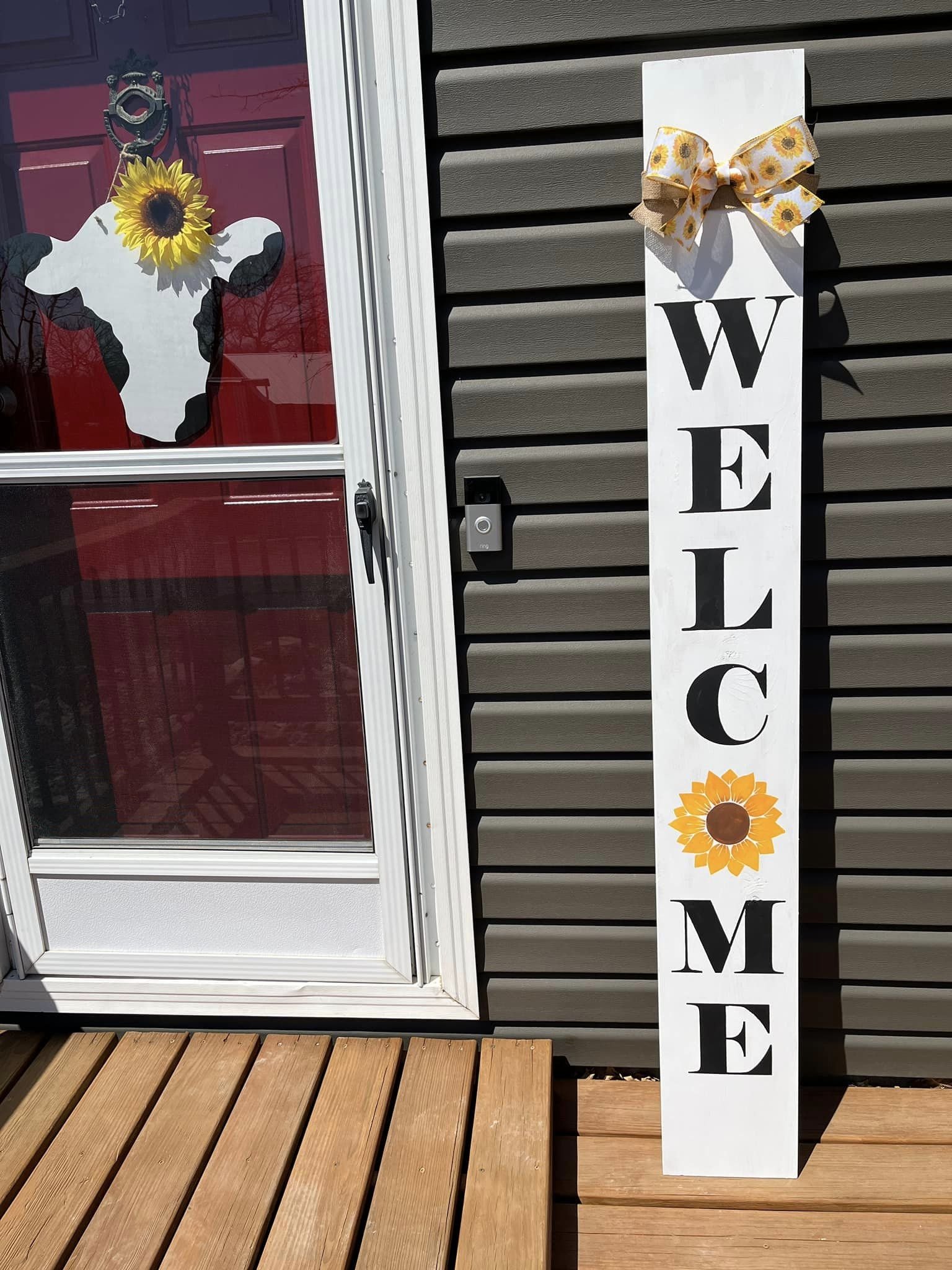 A white vertical welcome sign with a sunflower decoration at the top and a sunflower illustration in the middle, placed outside against a dark gray horizontal siding house. A door with a decal of a cow and a sunflower in its eye is also visible.