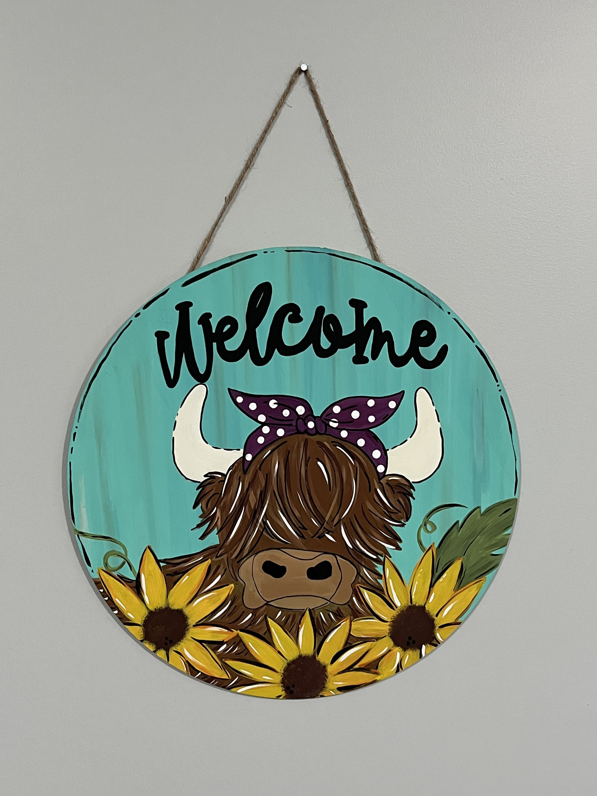 Decorative round sign with a teal background, featuring the word 'Welcome' at the top, and a cartoon of a Highland cow with long brown hair, white horns, and a purple polka-dotted bow, surrounded by yellow sunflowers.