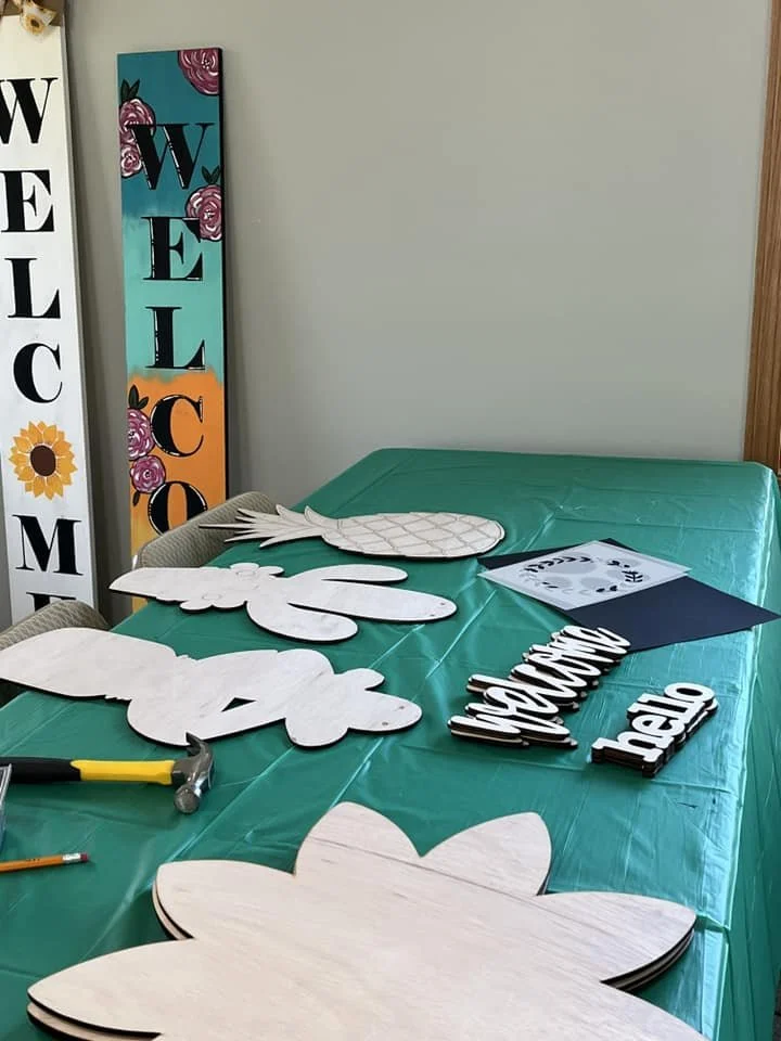 Worktable with laser-cut signs for a tropical or island-themed decoration, including a large pineapple, palm trees, and wood words saying "breathe" and "hello." Some tools are visible, and there are colorful signs on the wall that say "Welcome."