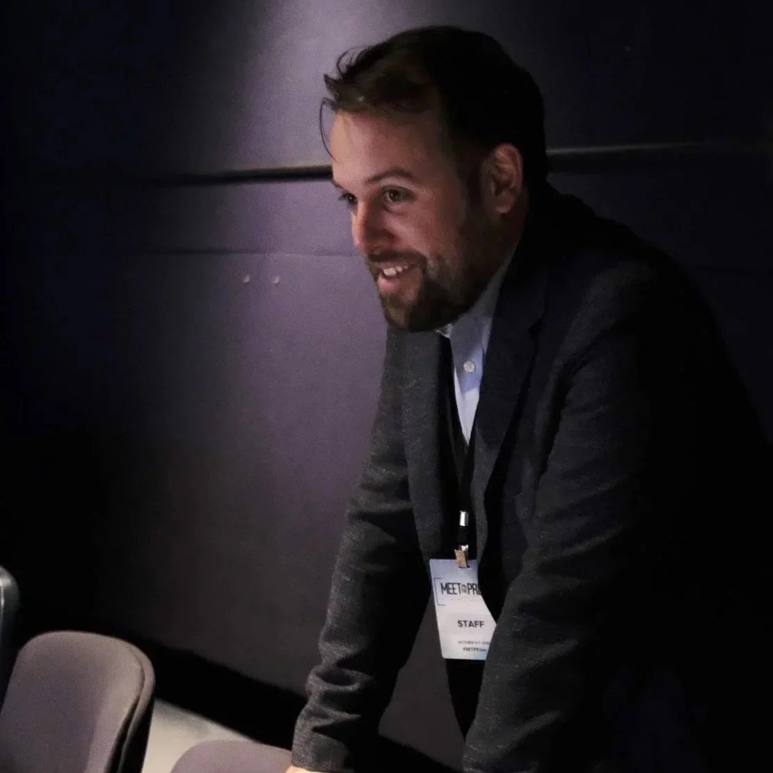 A man with a beard and short hair wearing a gray blazer and a staff badge, smiling and leaning forward in a dimly lit room with a black background.