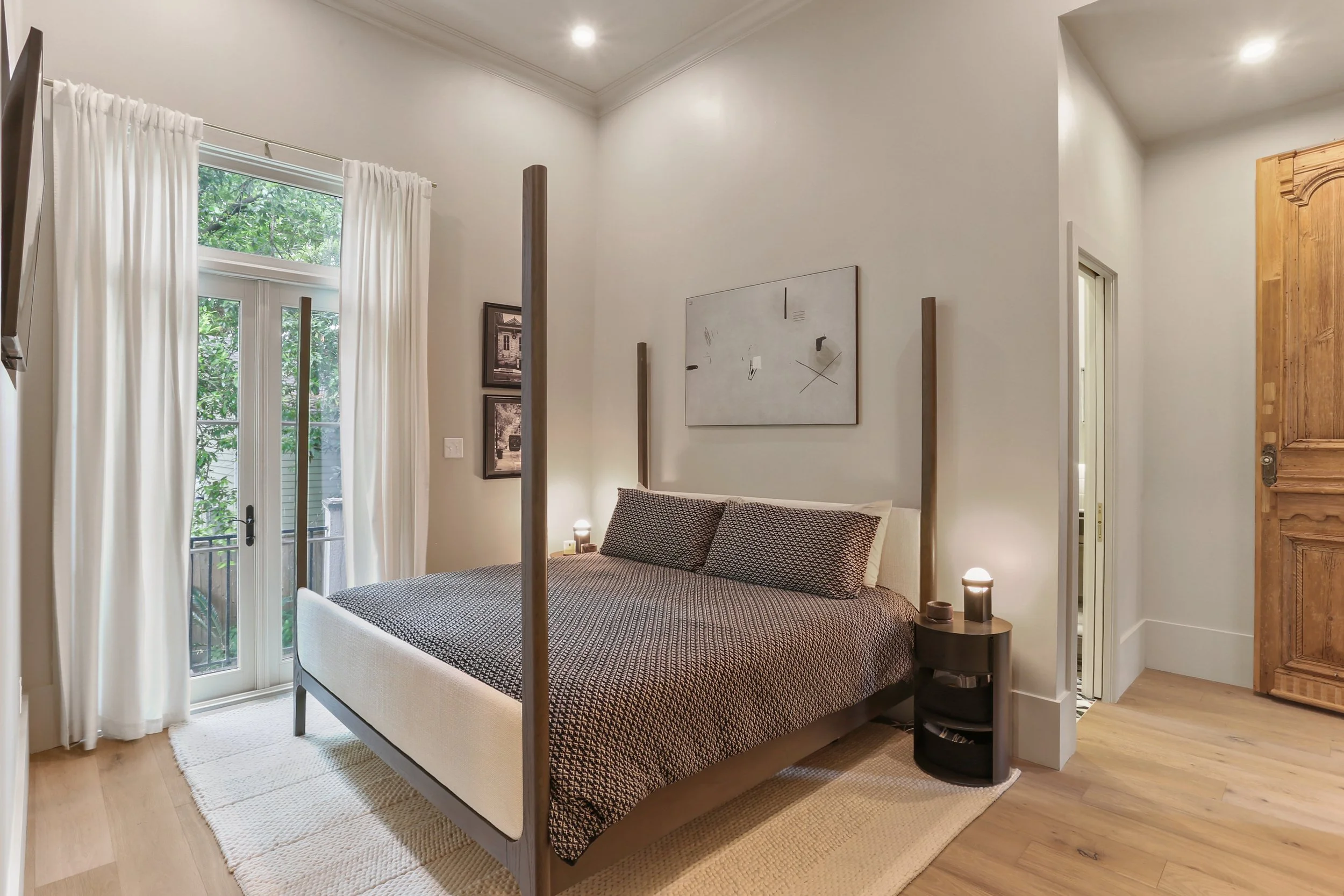 A modern bedroom with a wooden four-poster bed, neutral-colored bedding, a small round black bedside table with a lamp, white curtains on glass doors opening to a balcony, framed artwork on the walls, and wooden flooring.