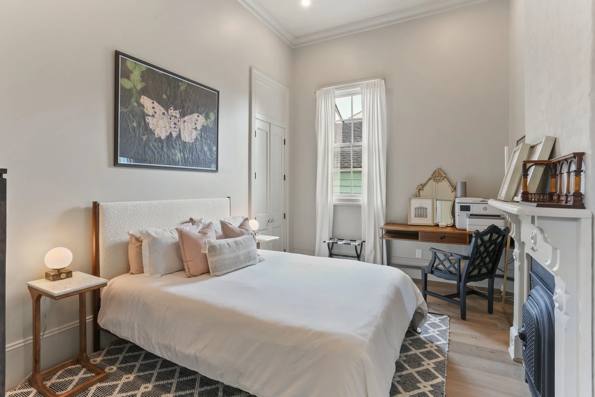 A bedroom with a white bed, pillows, nightstand lamps, framed butterfly artwork, a window with white curtains, a wooden desk with mirror and printer, black chair, and a white ornate fireplace.