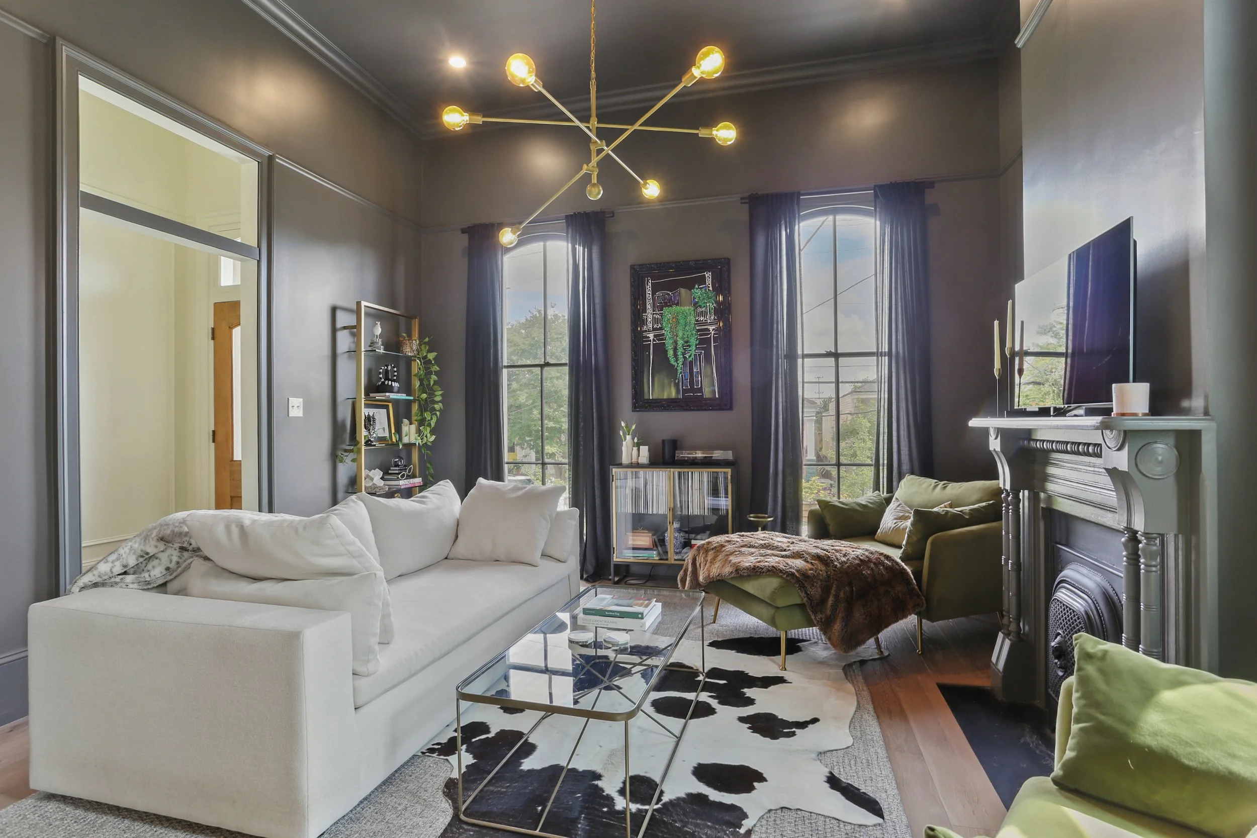 Living room with a white sofa, black and white cow print rug, green armchair with throw blanket, fireplace, flat-screen TV, and black curtains on large windows with trees outside. Modern chandelier hanging from ceiling.