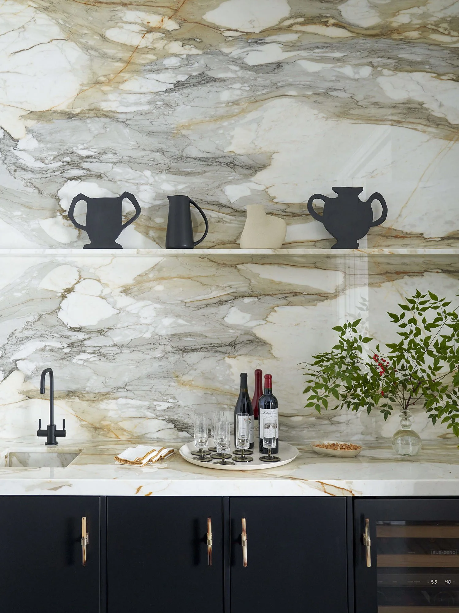Kitchen with marble backsplash and countertop, black sink and faucet, black cabinet doors with gold handles, decorative black vases, a tray with wine bottles and glasses, a bowl of snacks, and a vase with green leaves and red berries.