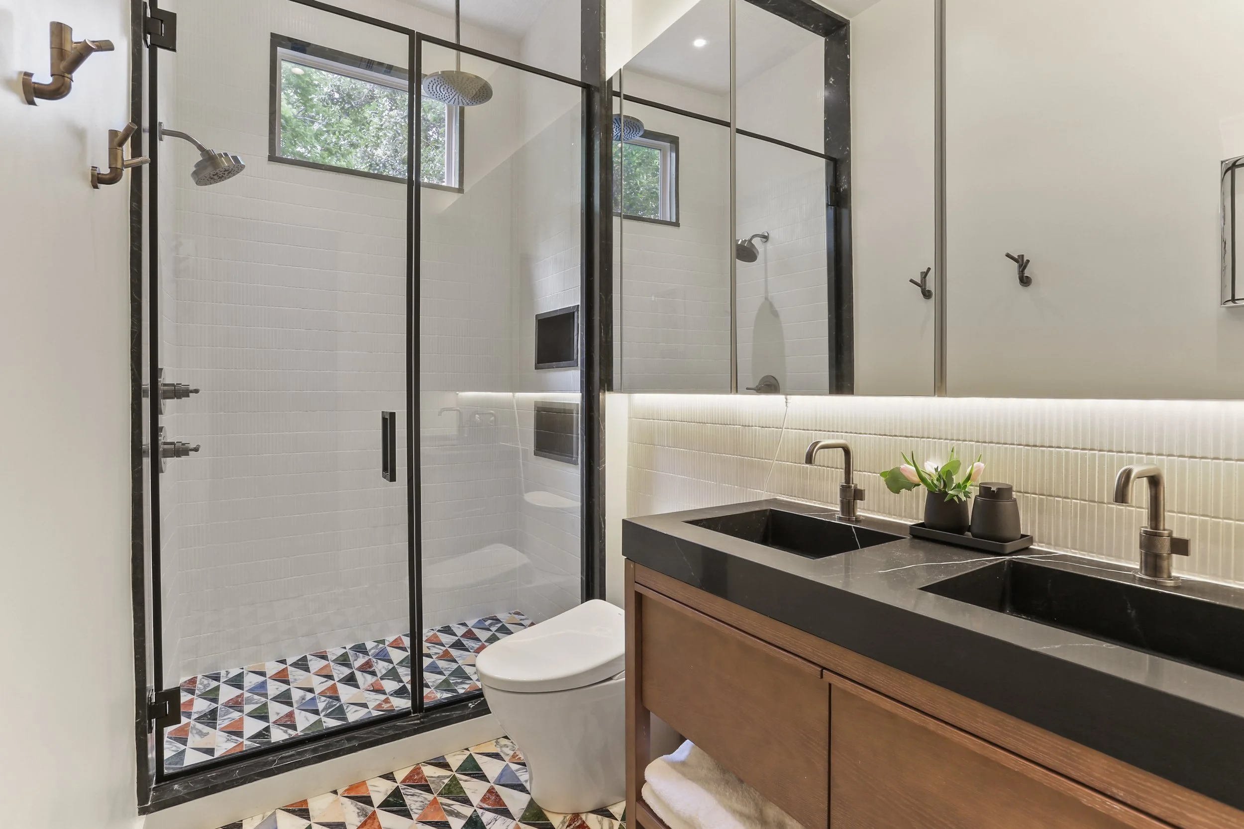 Modern bathroom with a separate glass-enclosed shower area, dual black sinks on a wooden vanity, a large mirror with backlighting, a white toilet, black fixtures, and geometric patterned tile floor.