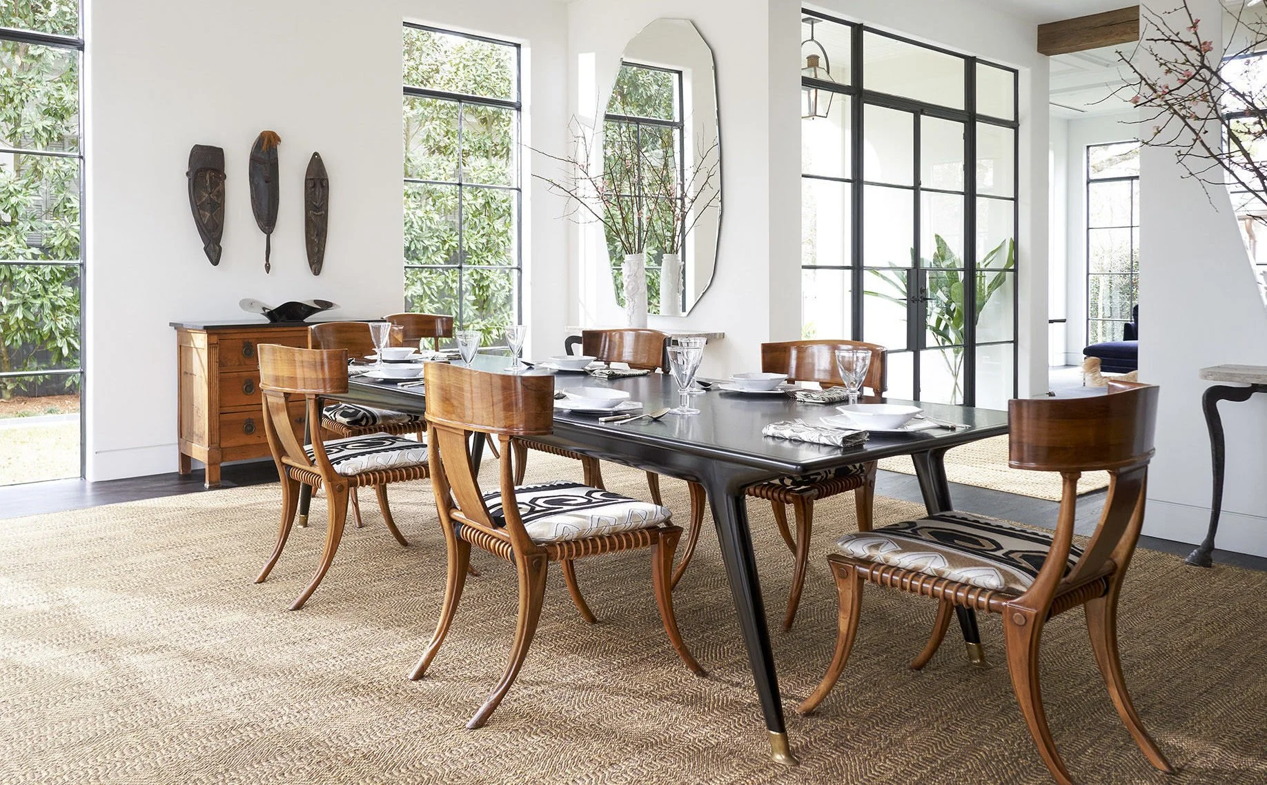 A modern dining room with a black table, wooden chairs, and large windows with black frames allowing natural light. The room has a beige rug, white walls, and decorative masks on the wall.