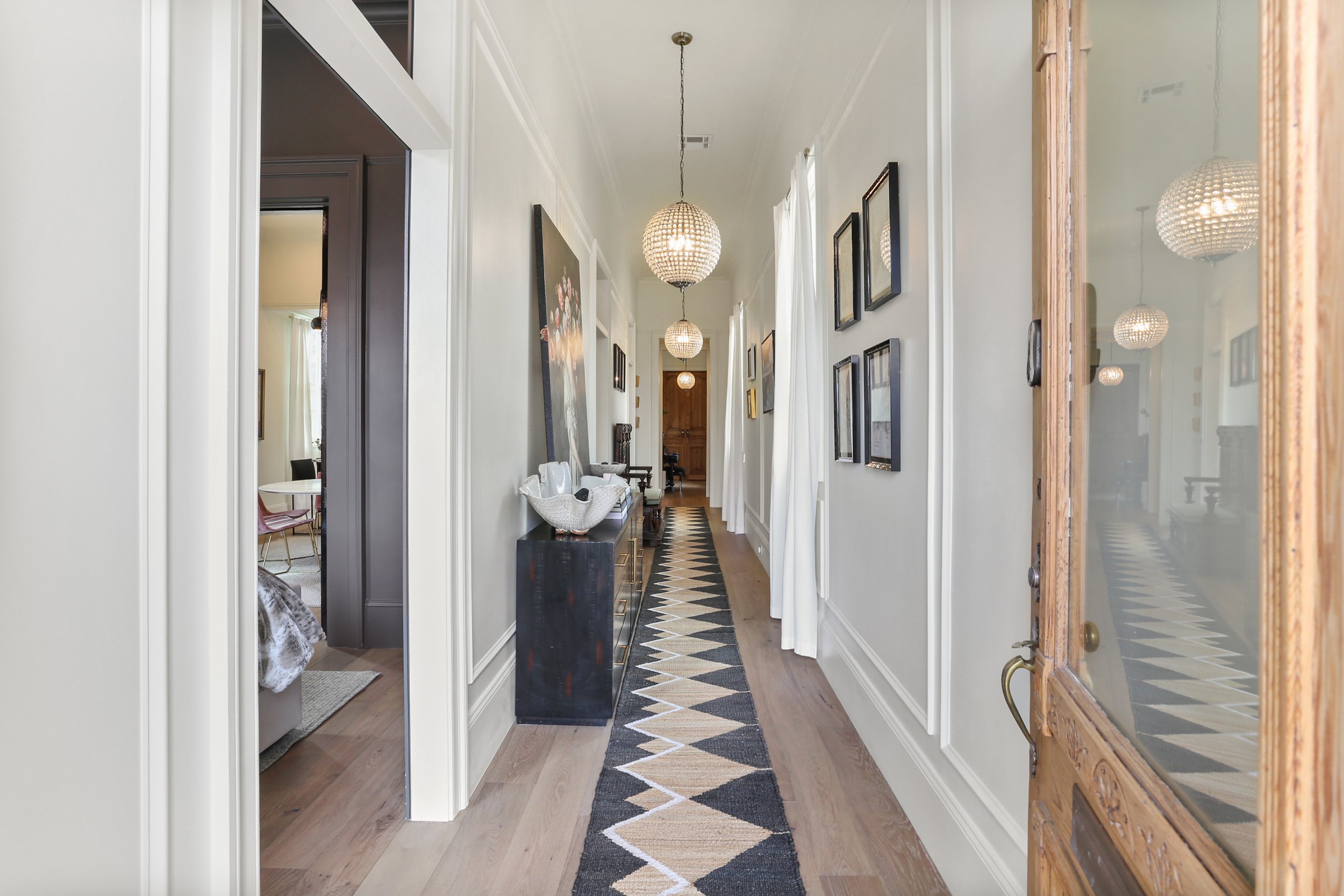 A hallway with light-colored walls, wooden flooring, and decorative wall moldings. Hanging spherical pendant lights line the ceiling. There is a patterned runner rug on the floor, and various framed pictures on the right wall. A black cabinet with decorative items is on the left side near the doorway to another room.