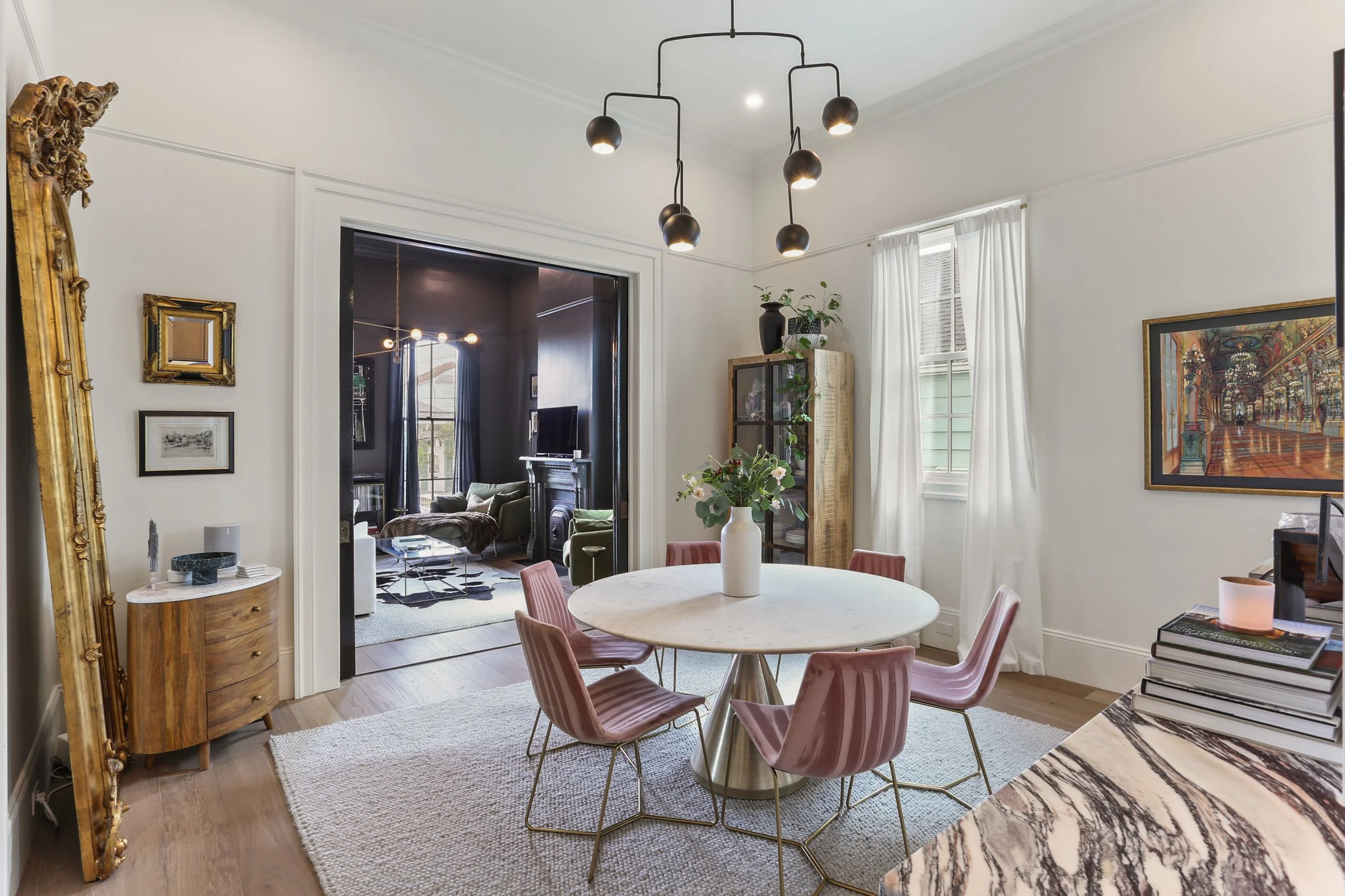 Dining room with round marble table, six pink velvet chairs, a white vase with flowers, wooden sideboard, artwork, and a view into the living room with green chairs and a fireplace.
