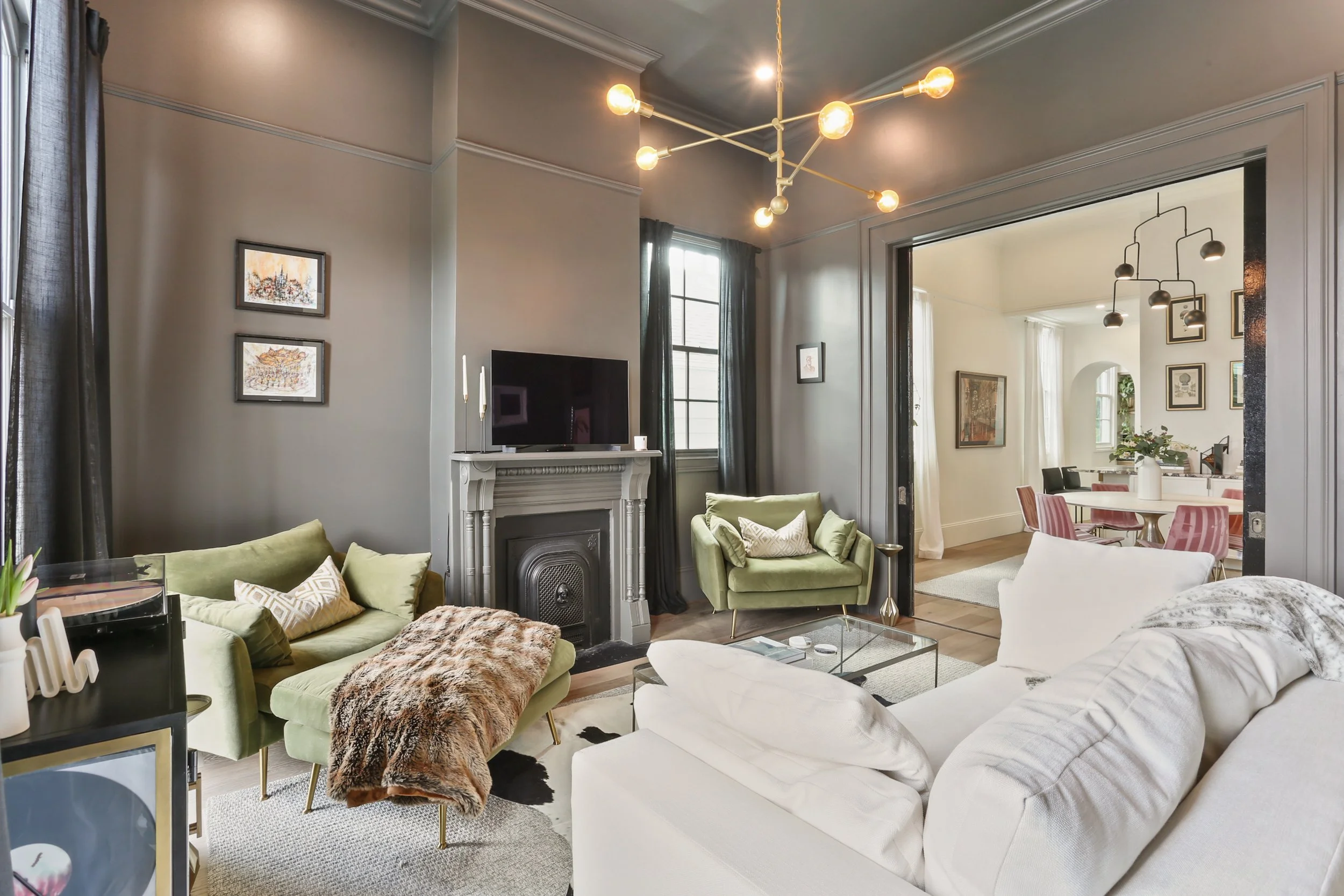 Living room with gray walls, green cushioned chairs, a white sofa, a modern chandelier, a fireplace with a TV on top, and a view into a dining area with pink chairs and wall art.