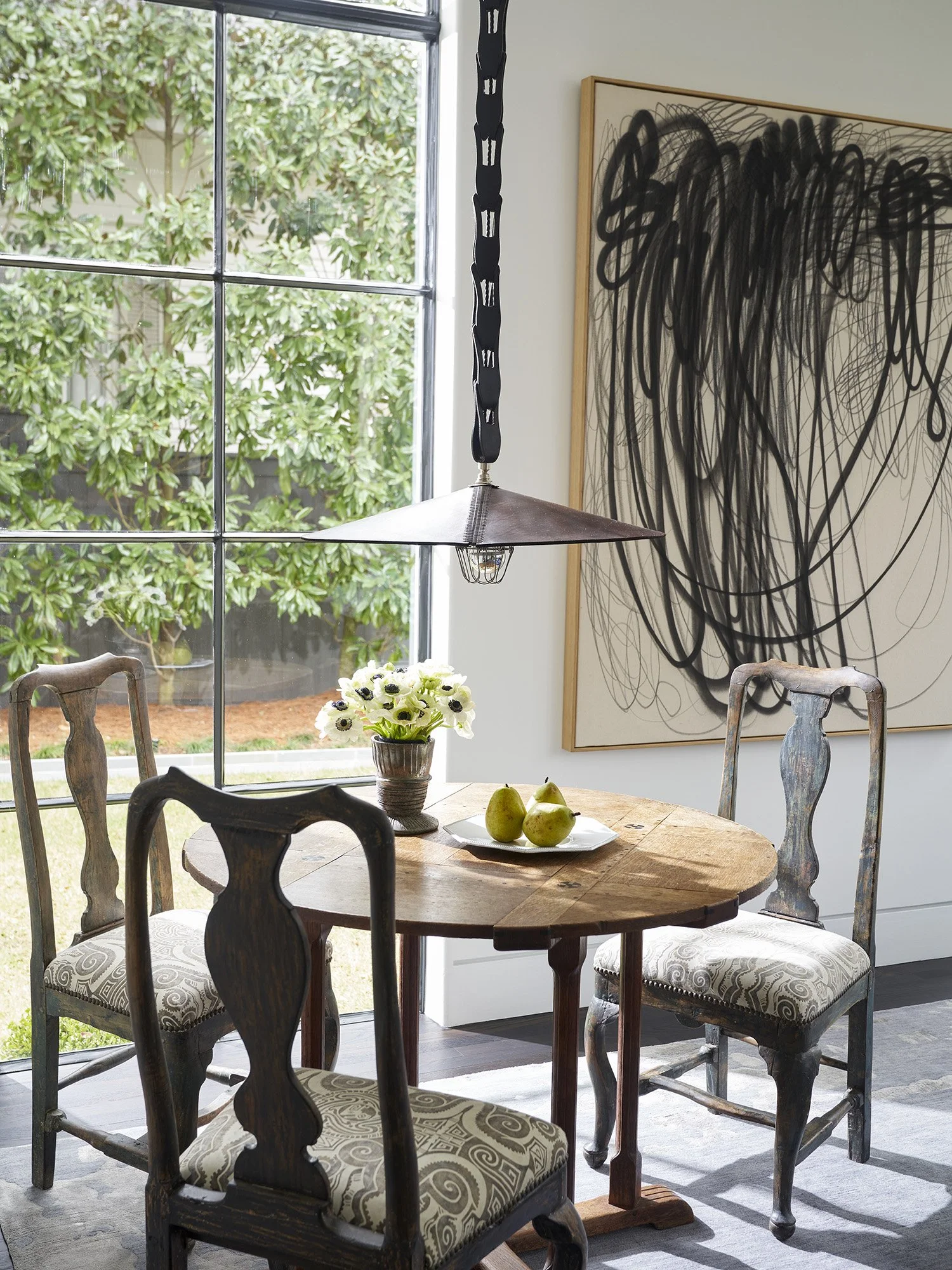 A round wooden dining table with three vintage-style chairs, a vase of white flowers, a plate of pears, a large black and white abstract artwork, and a black pendant light hanging above, in front of a glass door with greenery outside.