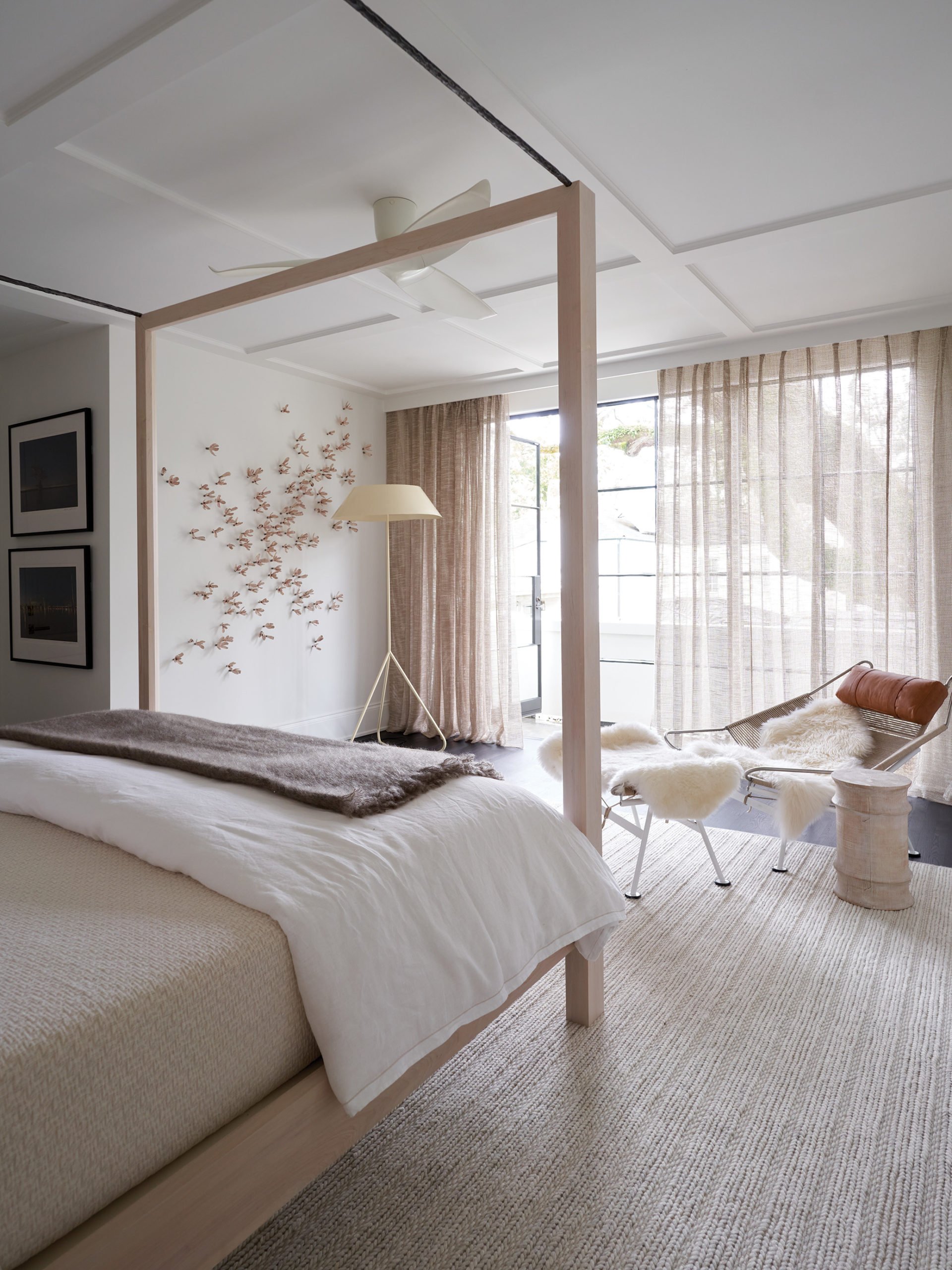 Bright bedroom with a canopy bed, beige bedding, and a brown blanket. A hanging chair with white fur and a small table nearby. Large windows with sheer curtains allowing sunlight. Wall art featuring birds and framed pictures.