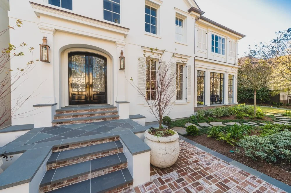 Front entrance of a large white house with black doors, brick steps, and a landscaped garden with trees and plants.
