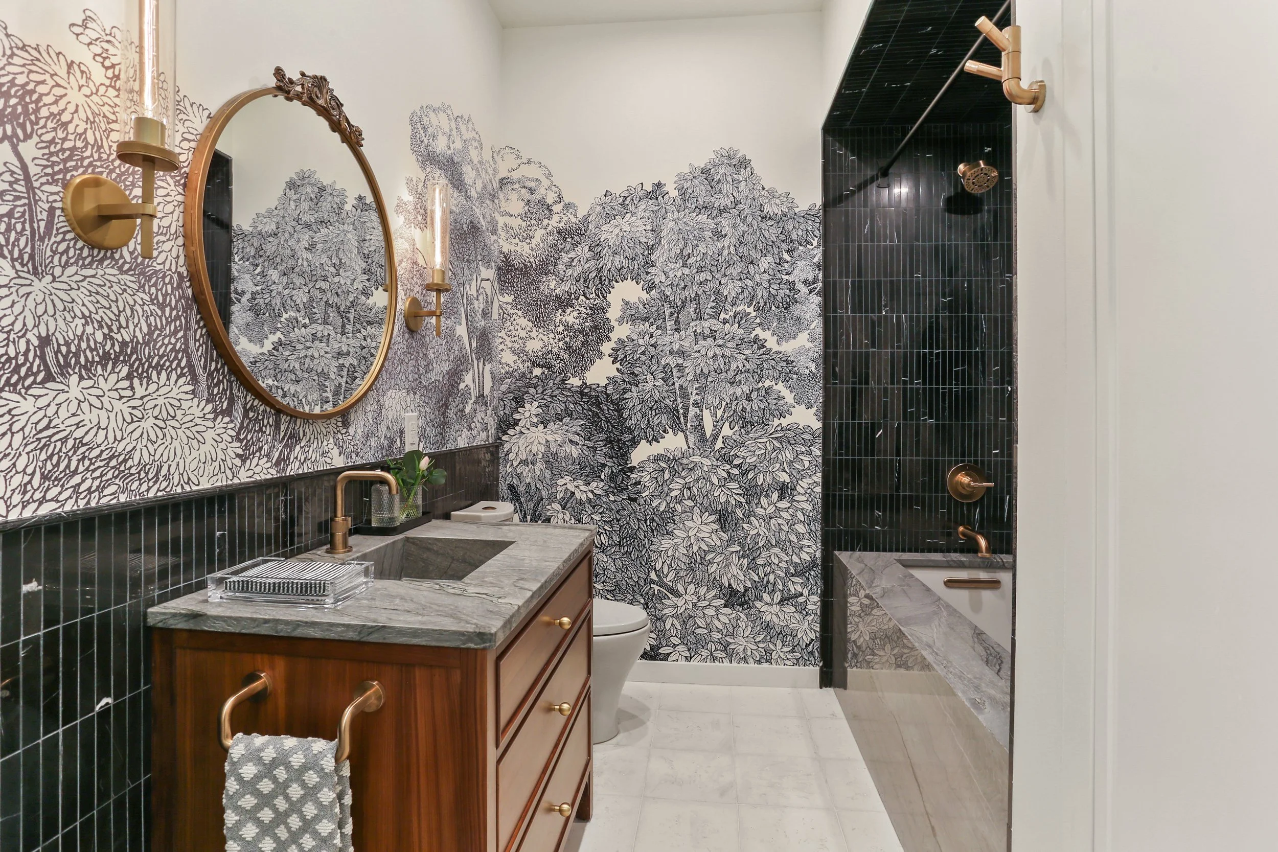 Modern bathroom with black and white botanical mural, wooden vanity with gray marble countertop, round mirror, candle sconces, and black tiled shower area with black fixtures.